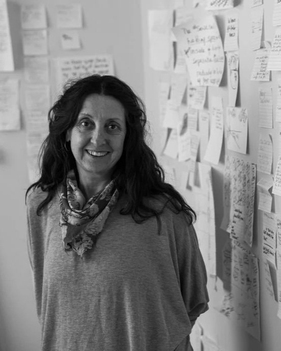 A woman with curly hair smiling and standing in front of a wall covered with notes and papers.