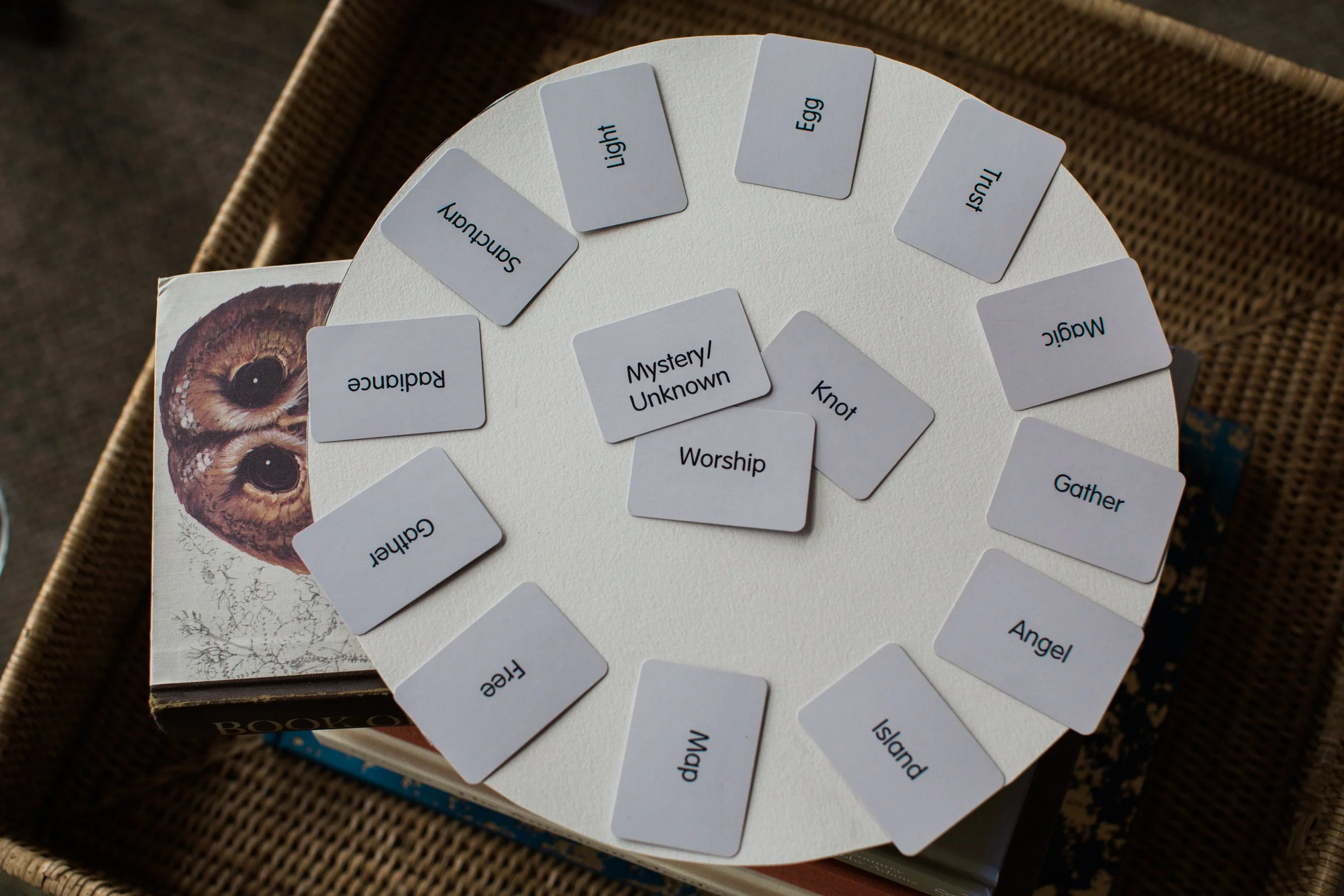 A circular game board with flash cards containing words related to love and relationships, such as 'Secret', 'Mind', 'Knot', and 'Mystery/Unknown'. The board features an image of owl eyes and is placed in a woven basket.