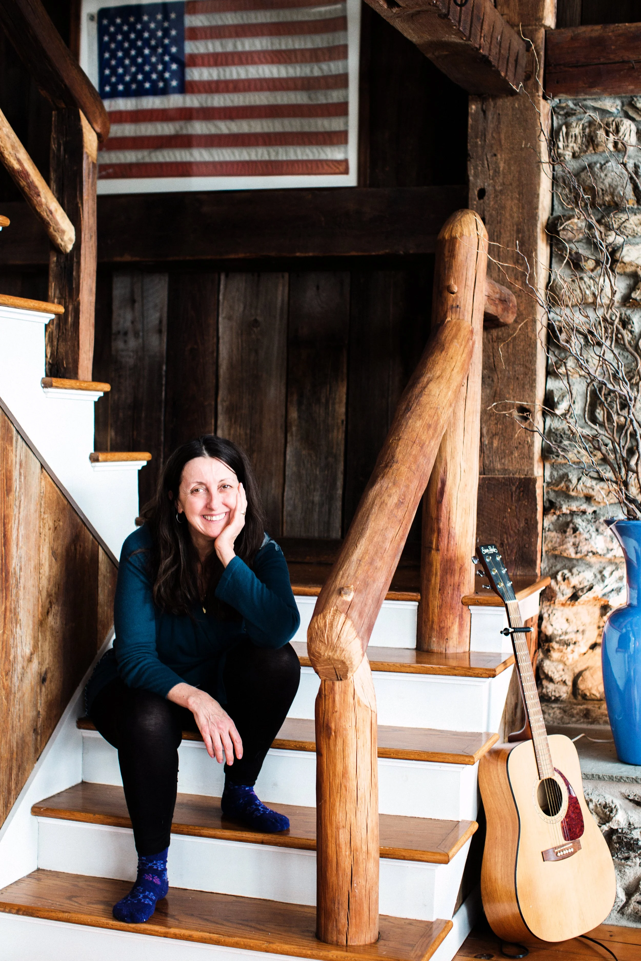 A woman sitting on a wooden staircase in a rustic home. She has long dark hair, is wearing a blue sweater and black jeans, and is smiling while resting her face in one hand. An acoustic guitar leans against the wall beside her. An American flag hangs on the wall in the background.