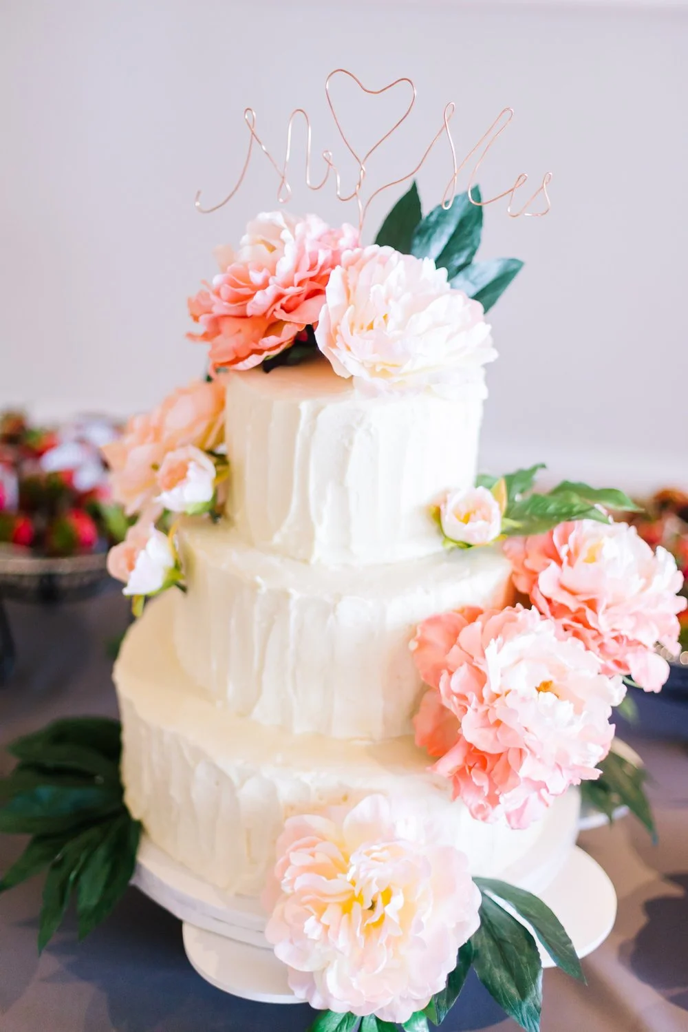 Three-tier white wedding cake decorated with pink flowers and a wire topper reading Mr / Mrs.