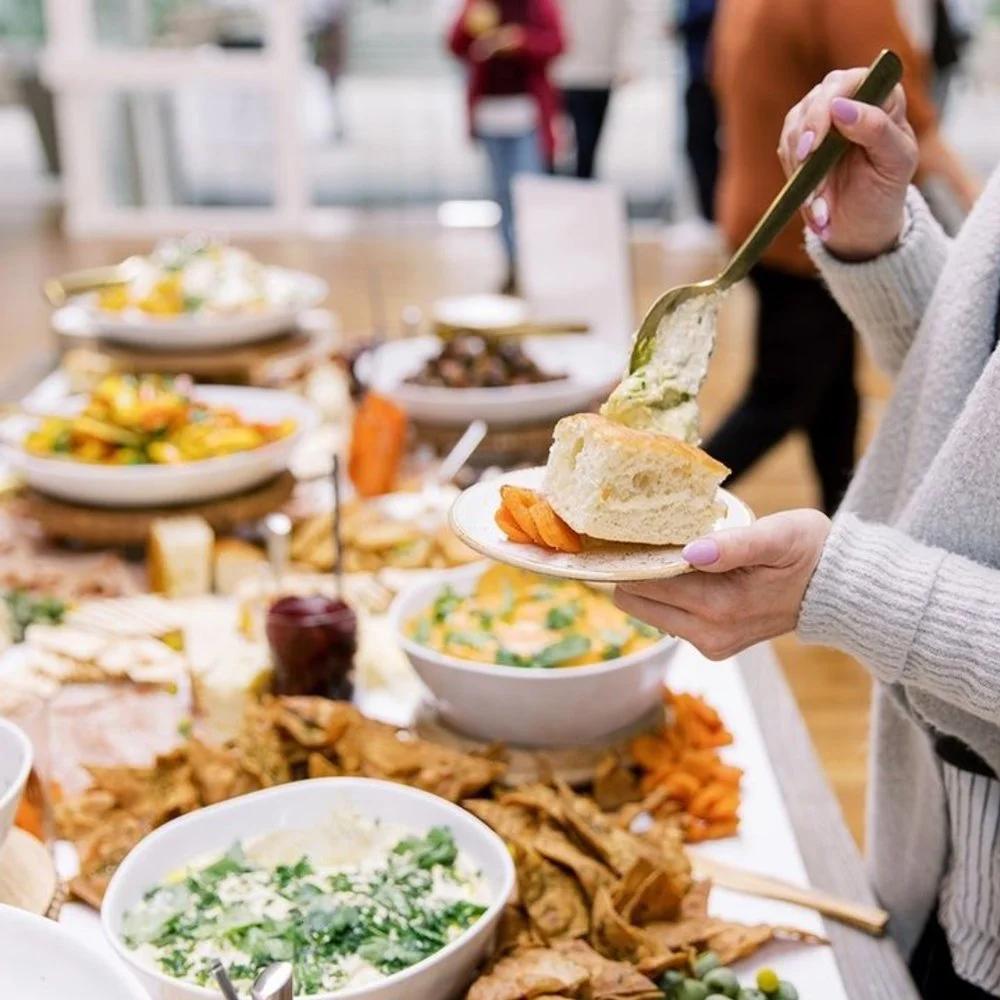 Guest at a social event dolloping dip on bread slice at a buffet offering various dips and chips.