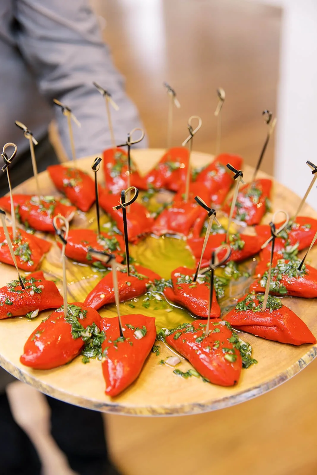Red stuffed peppers on a wooden platter with herbs and olive oil, each secured with a decorative pick.