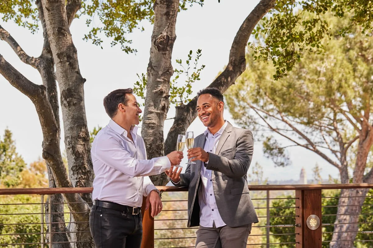Two men in business attire toasting with champagne glasses while leaning on the railing of the Tree Deck at The Quad Conference Center.