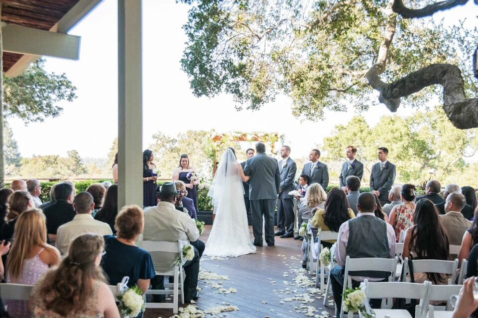 Outdoor wedding ceremony on the Tree Deck at The Quad Conference Center with bride and groom in front, bridesmaids and groomsmen on each side, and guests seated behind them.