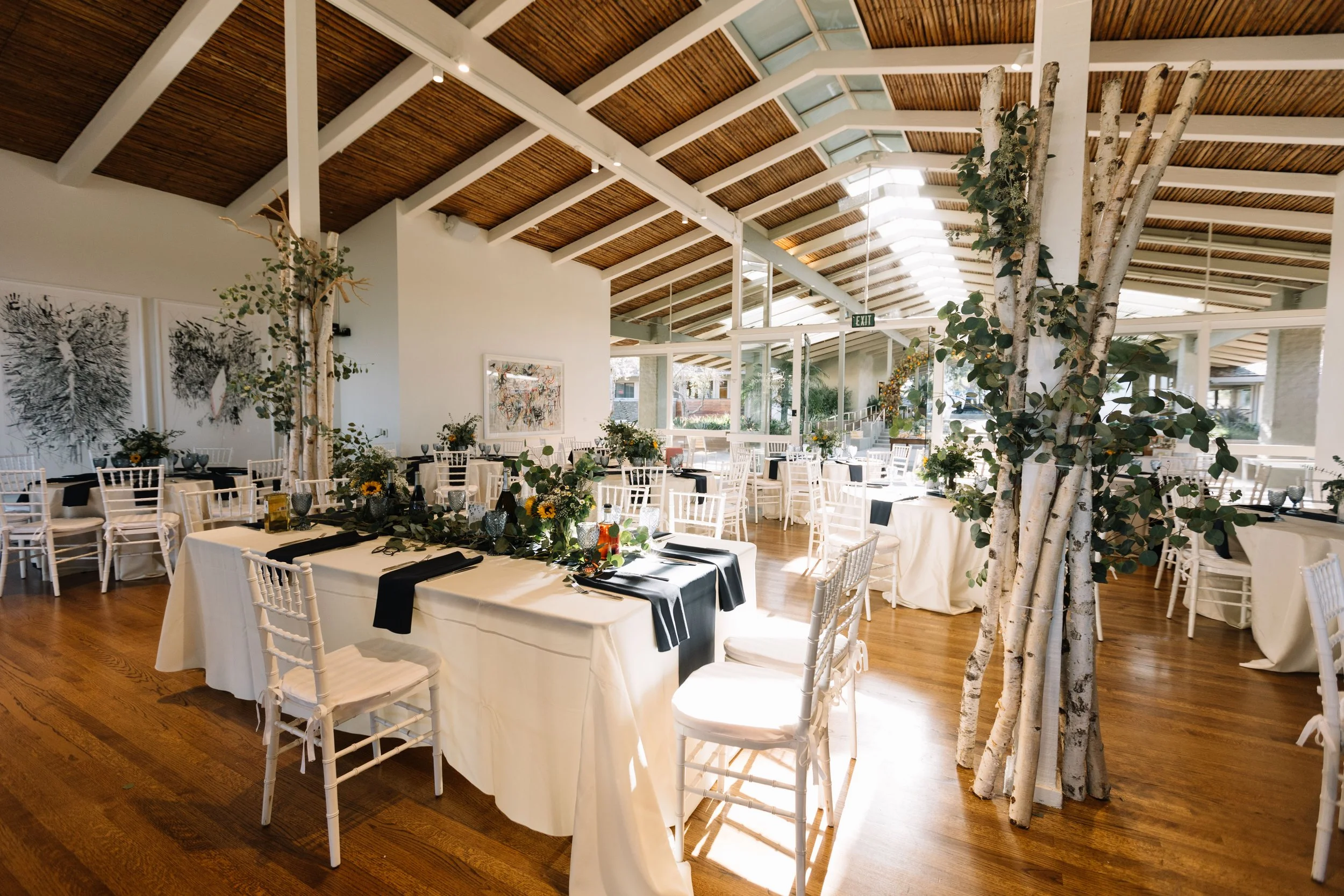 Wedding reception in the sun-lit Quad Conference Center with white chairs, tables, floral centerpieces, and wooden ceiling beams.