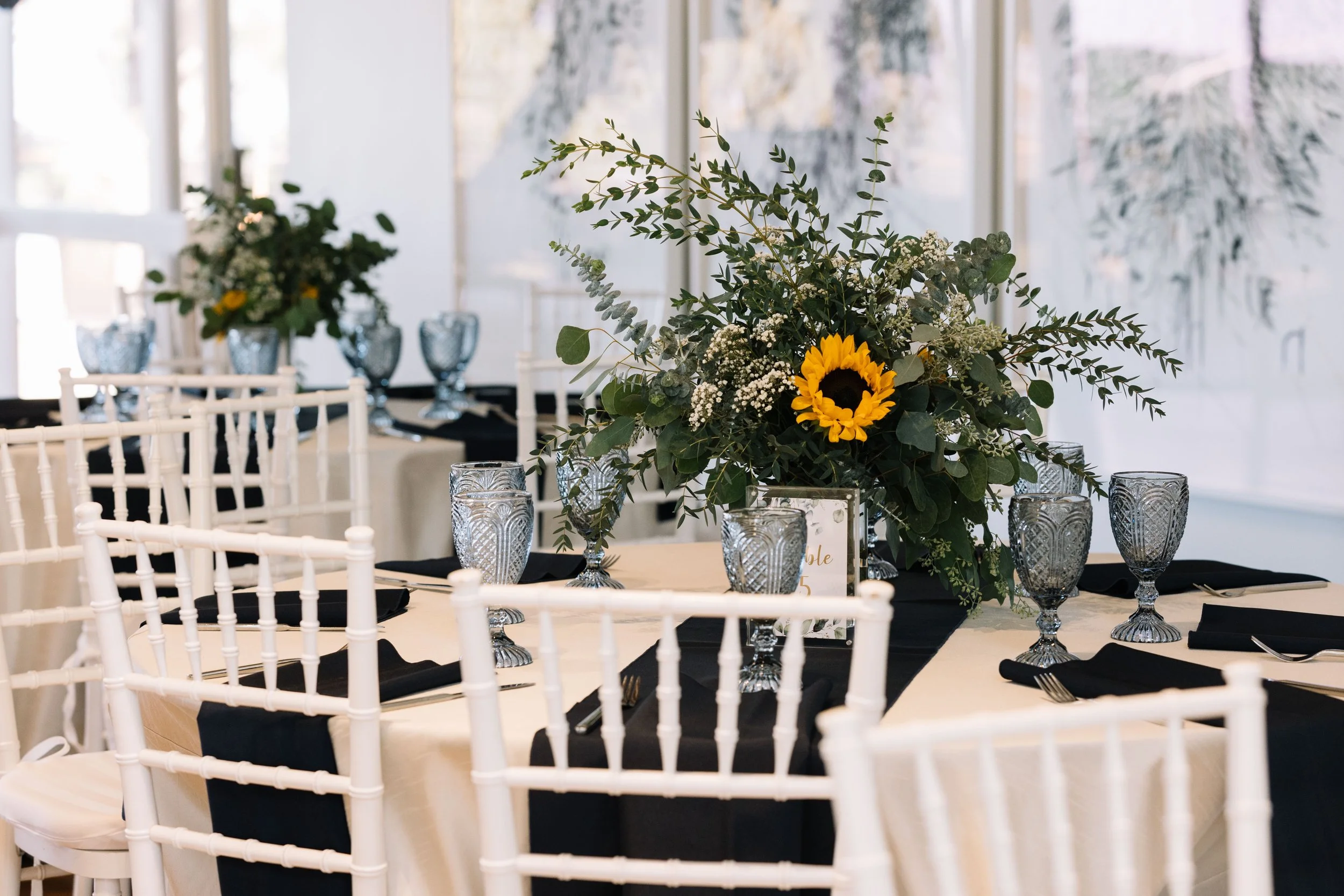 Elegant table setup for a wedding reception with white chairs, black napkins, decorative glasses, and sunflower centerpiece at The Quad Conference Center.