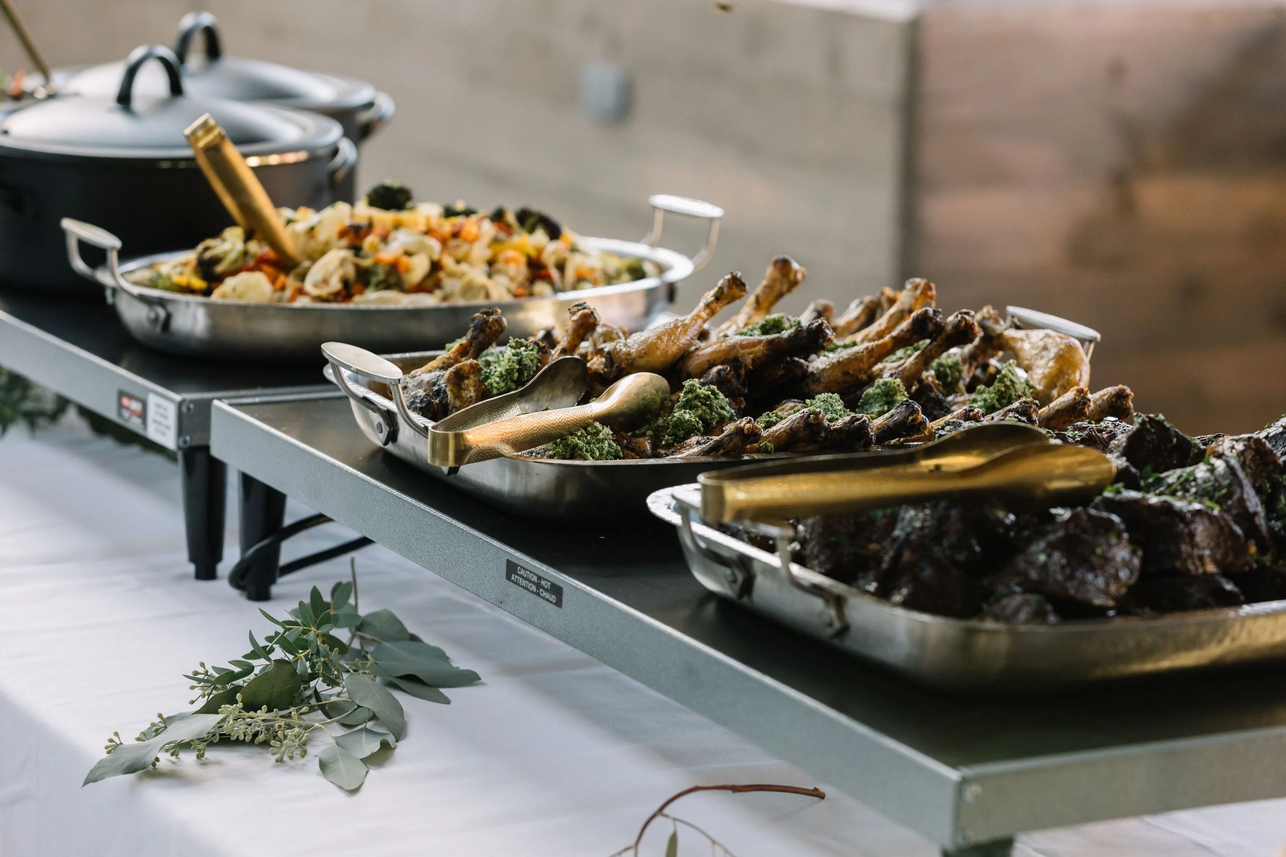 Buffet setup with trays of cooked chicken drumsticks and mixed vegetables on a serving table.