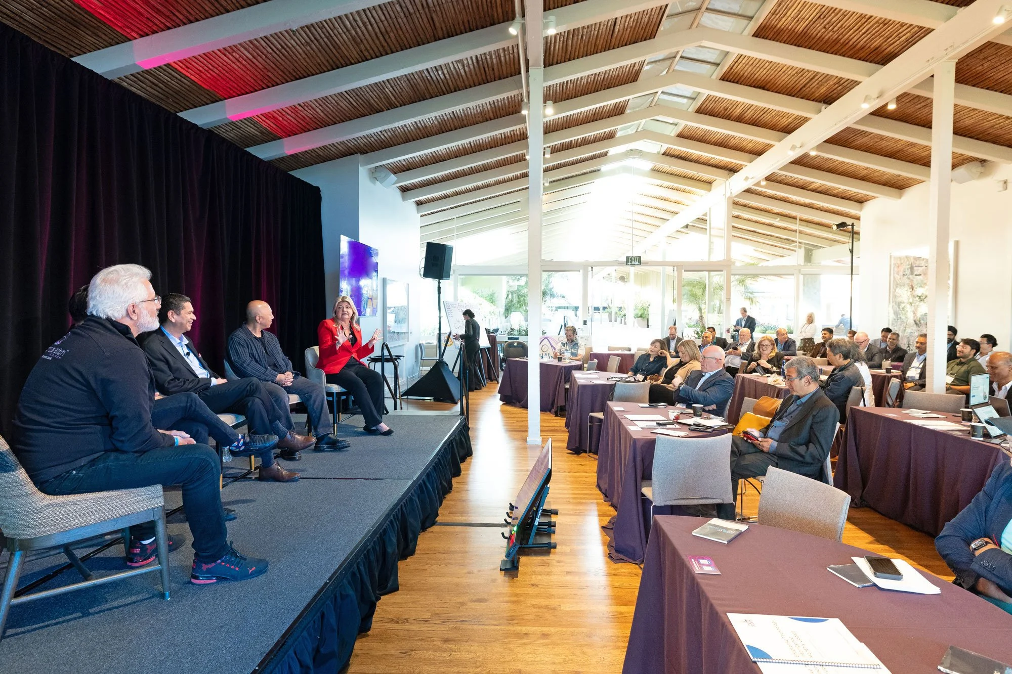 Panel of five speakers on stage at The Quad Conference Center with corporate event audience seated at tables listening.