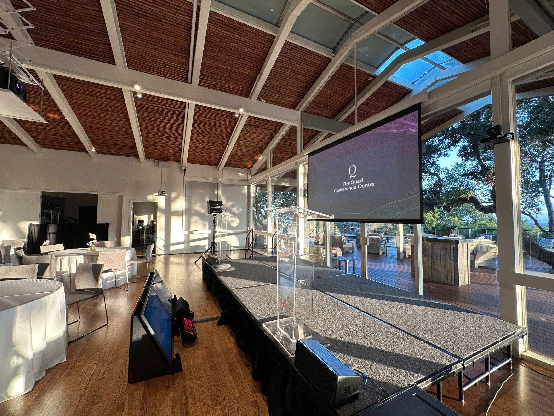 The stage at The Quad Conference Center set for a corporate event with a large projector screen under a ceiling with wooden panels with a view of the Tree Deck through the windows behind the screen.