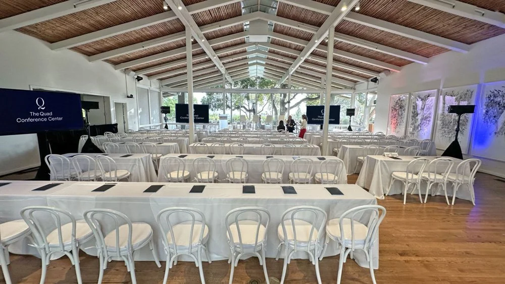 The Quad Conference Center set for a corporate event with rows of long tables covered in white tablecloths, white chairs, and multiple screen monitors under a ceiling with wooden panels.