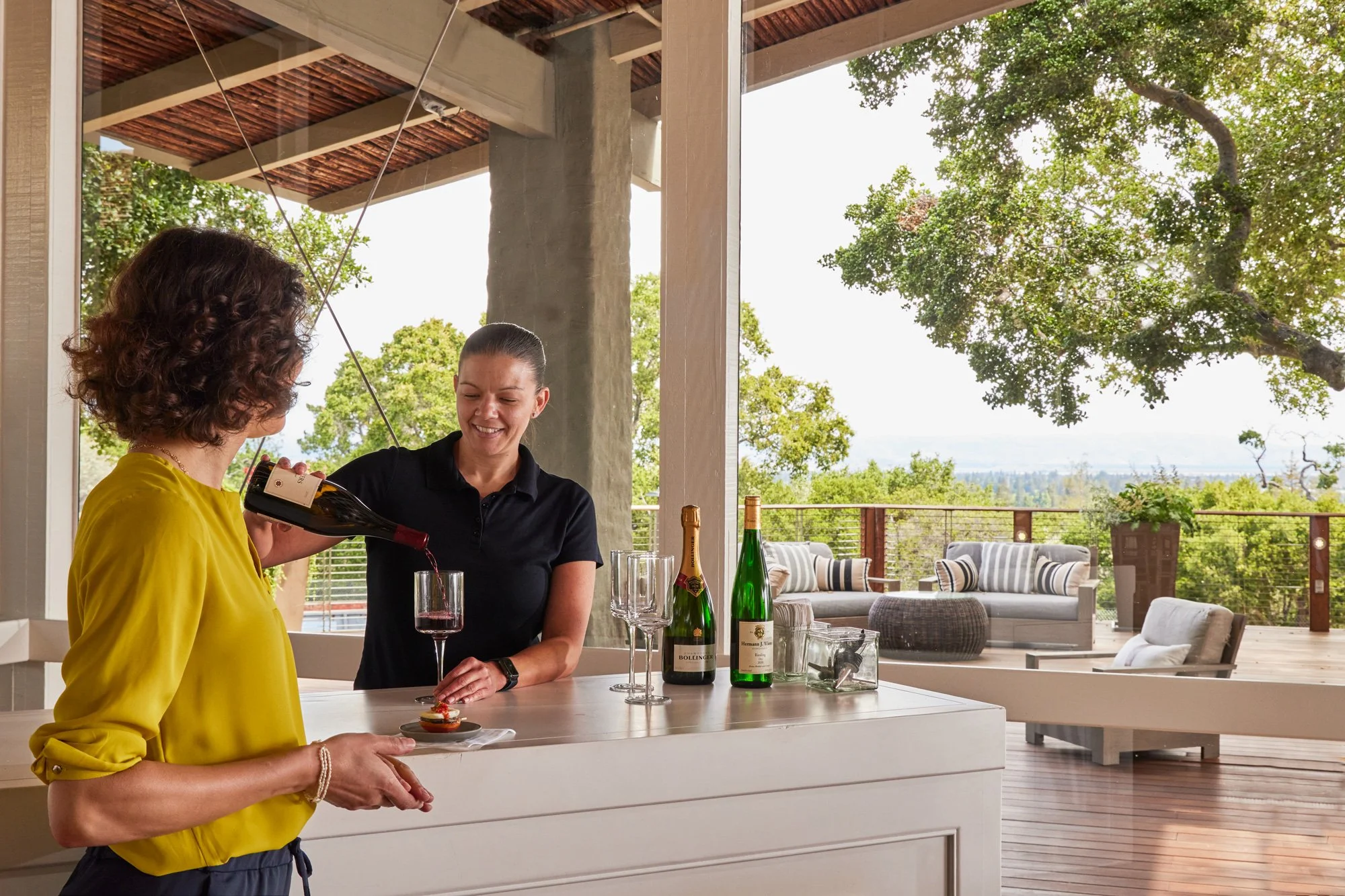 A bartender pours wine into a glass for a woman in The Quad Conference Center with comfortable couch seating arranged on the Tree Deck behind them.