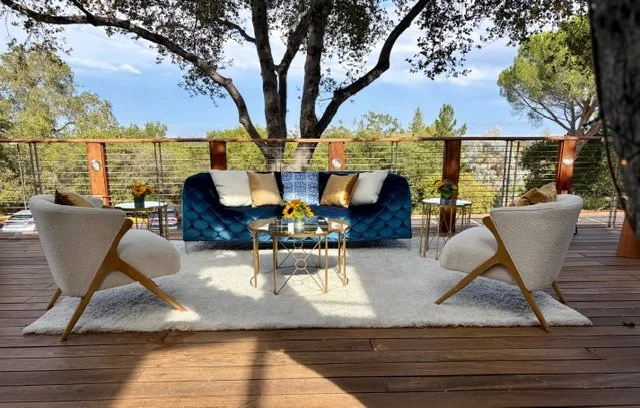 Outdoor seating on the Tree Deck at The Quad Conference Center with a blue sofa, two white chairs, gold-accented tables, and a white rug.