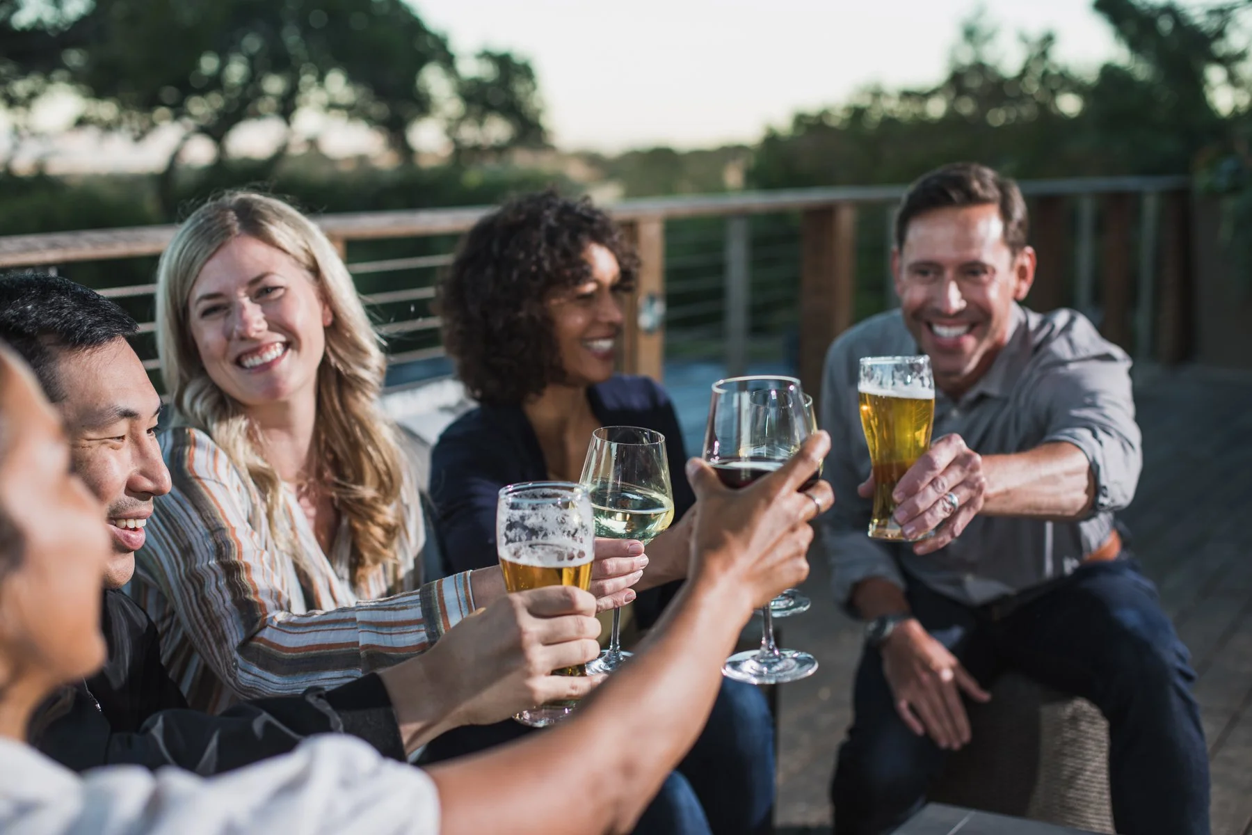 A group of diverse friends smiling and clinking glasses of beer and wine on the Tree Deck at The Quad Conference Center.