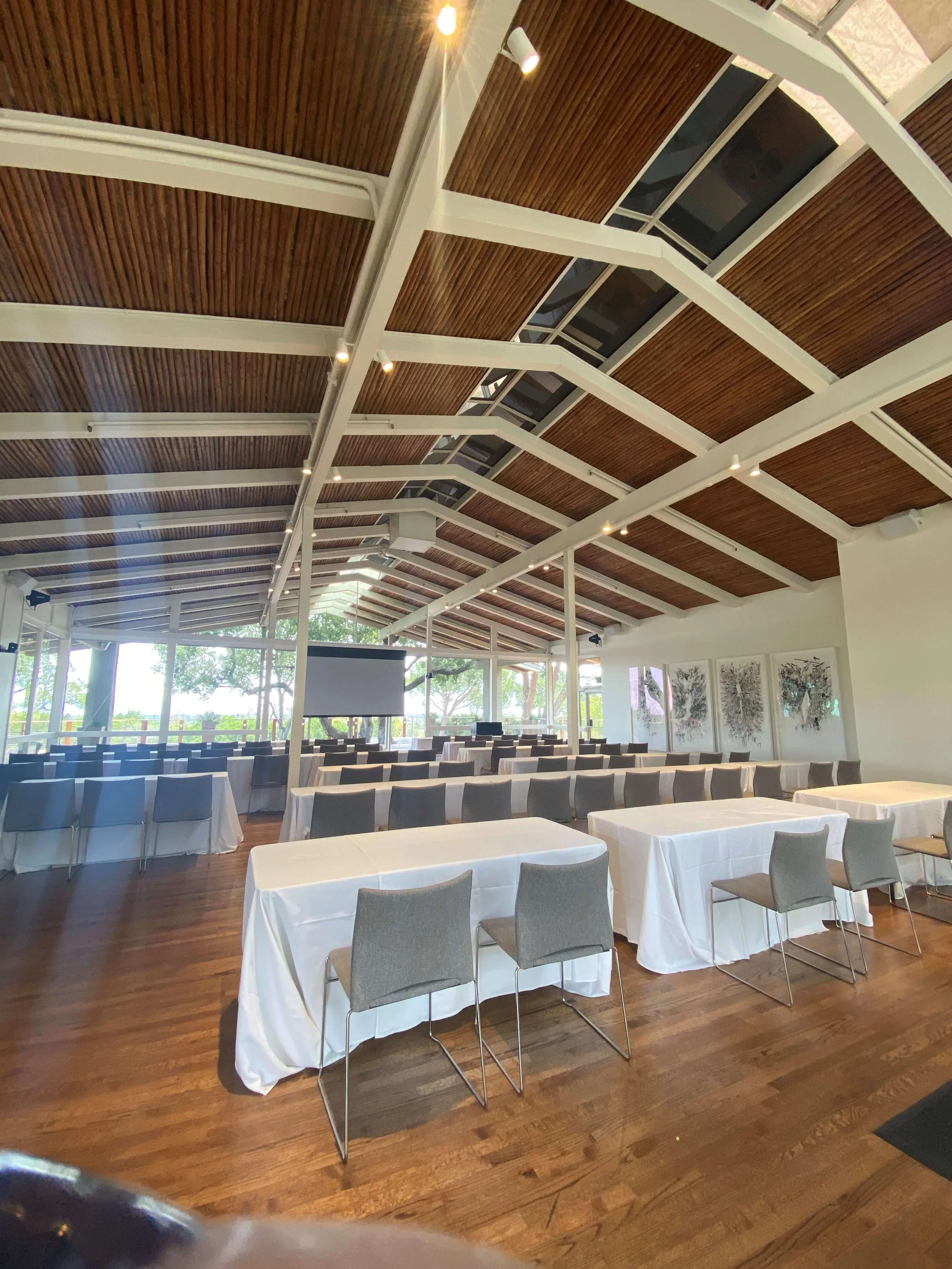 The atrium at The Quad Conference Center set for a corporate event with rows of tables covered in white tablecloths, gray chairs, and a projector screen under a ceiling with wooden panels.