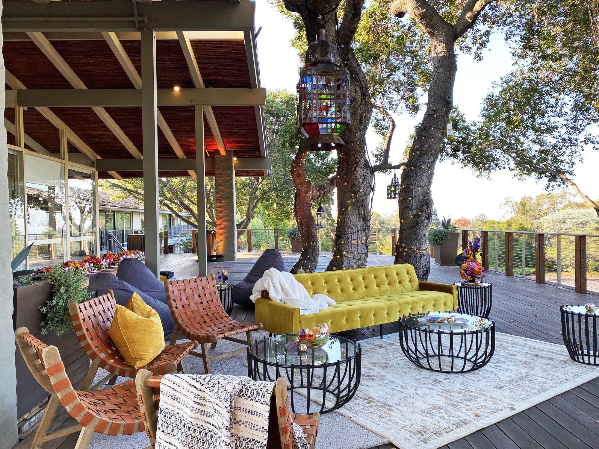 An inviting outdoor seating arrangement with a yellow couch, wicker chairs, glass tables, and string lights set up on the Tree Deck at The Quad Conference Center.
