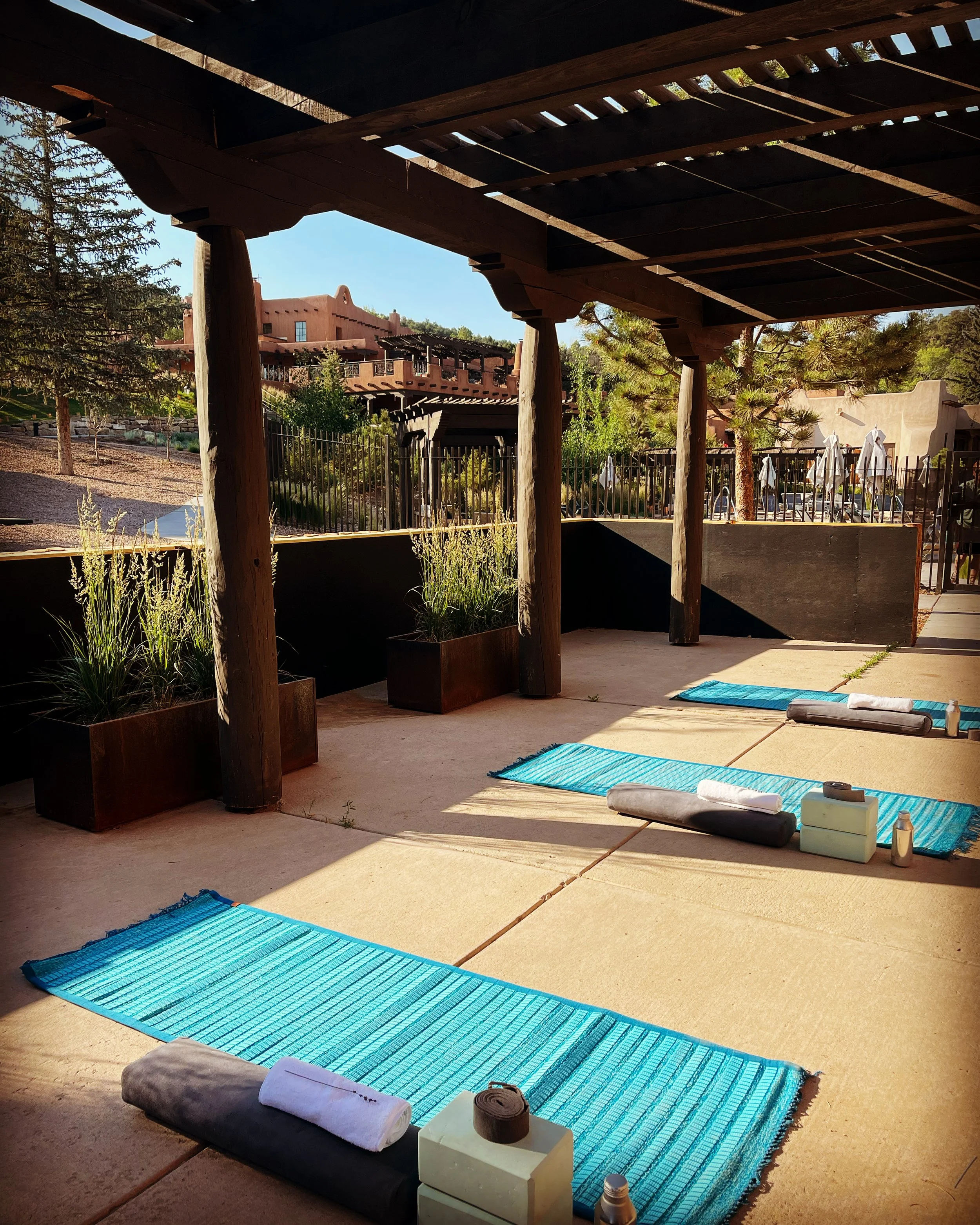 Yoga mats and props arranged outdoors under a wooden pergola, with a building and trees in the background.