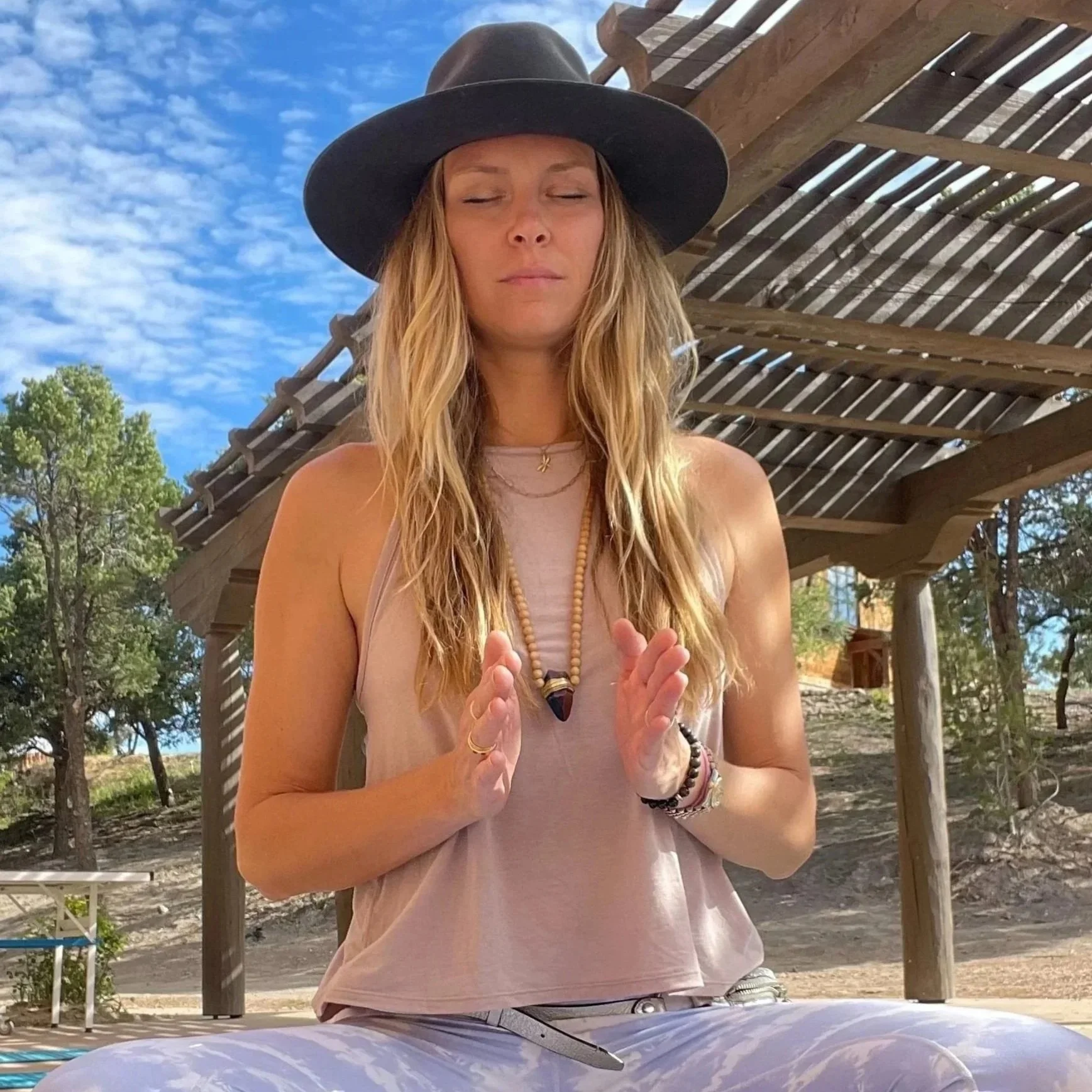 A woman with long blonde hair, wearing a large black wide-brimmed hat and a light pink sleeveless top, sitting outdoors on a sunny day with her eyes closed and hands in a prayer position. There are trees and a wooden structure in the background.