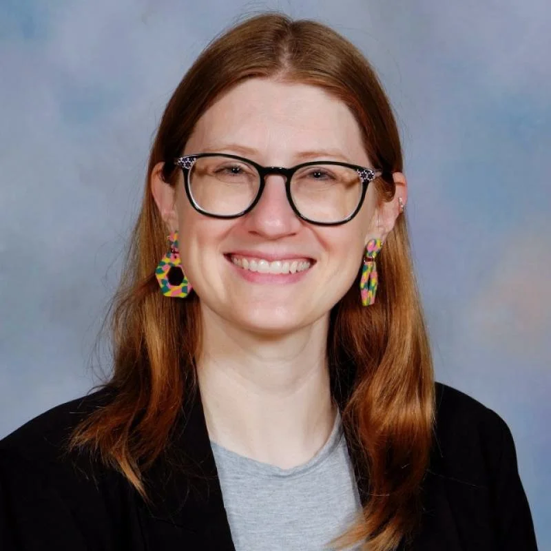 Headshot of a smiling woman with red hair, large black glasses with decorative accents, colorful earrings, wearing a gray shirt and black blazer, against a soft blue background.
