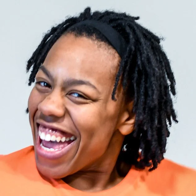 Close-up of a woman with dark, textured hair, wearing an orange top, smiling and winking.
