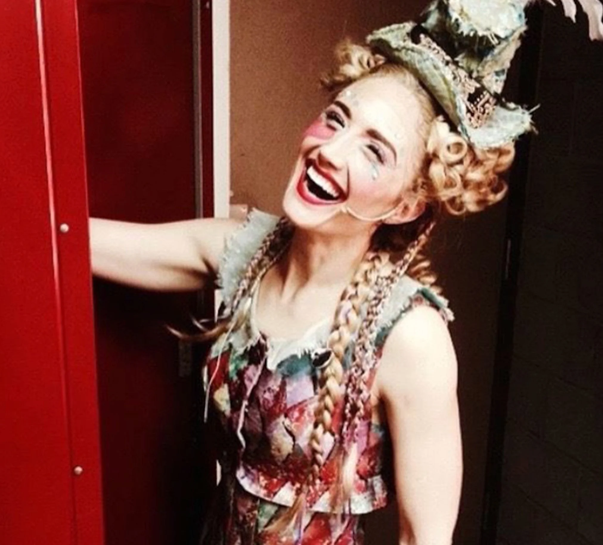 A woman with curly hair and braids, wearing a colorful dress and a headscarf, is smiling and leaning into a red locker.