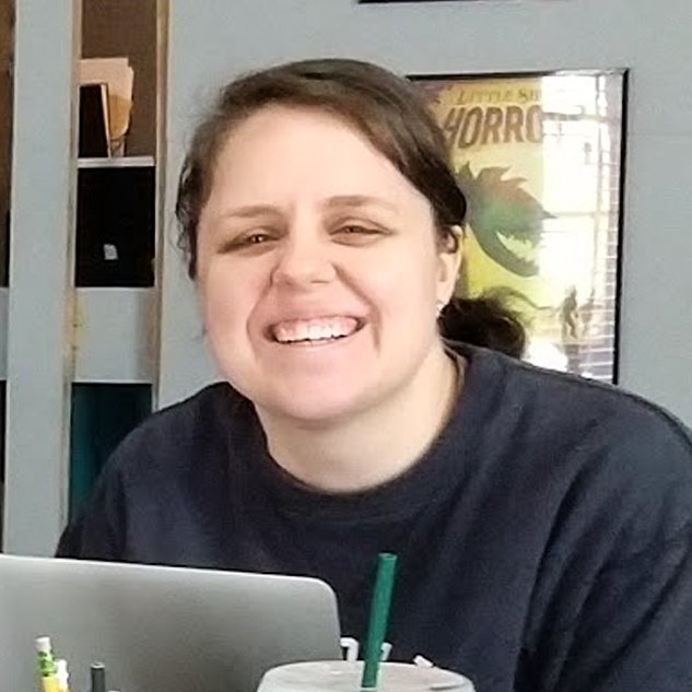 A woman with brown hair smiling at a desk with a laptop and a drink in a cup with a straw in front of her, sitting in a room with shelves and a framed poster on the wall.