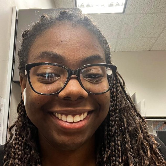 A young Black woman with glasses and braided hair smiling at the camera in an office setting.