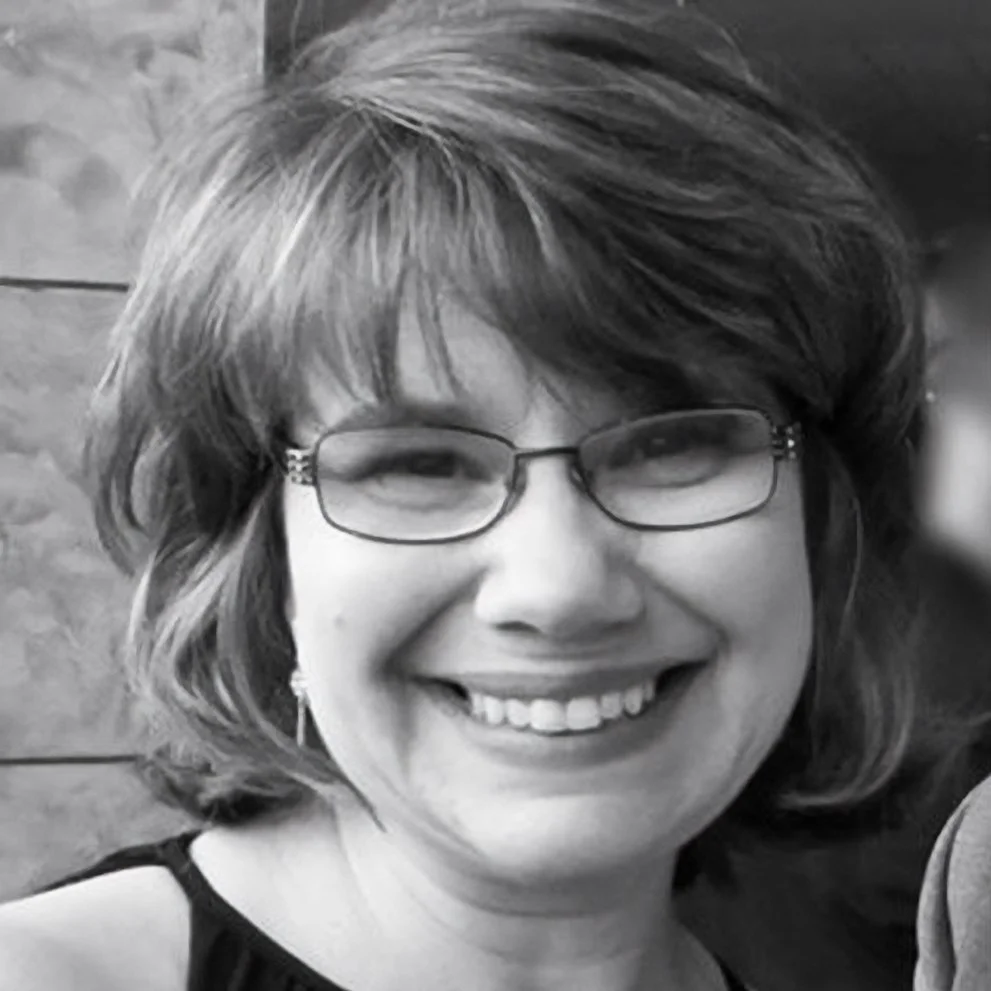 A smiling woman with glasses, short wavy hair, and earrings.