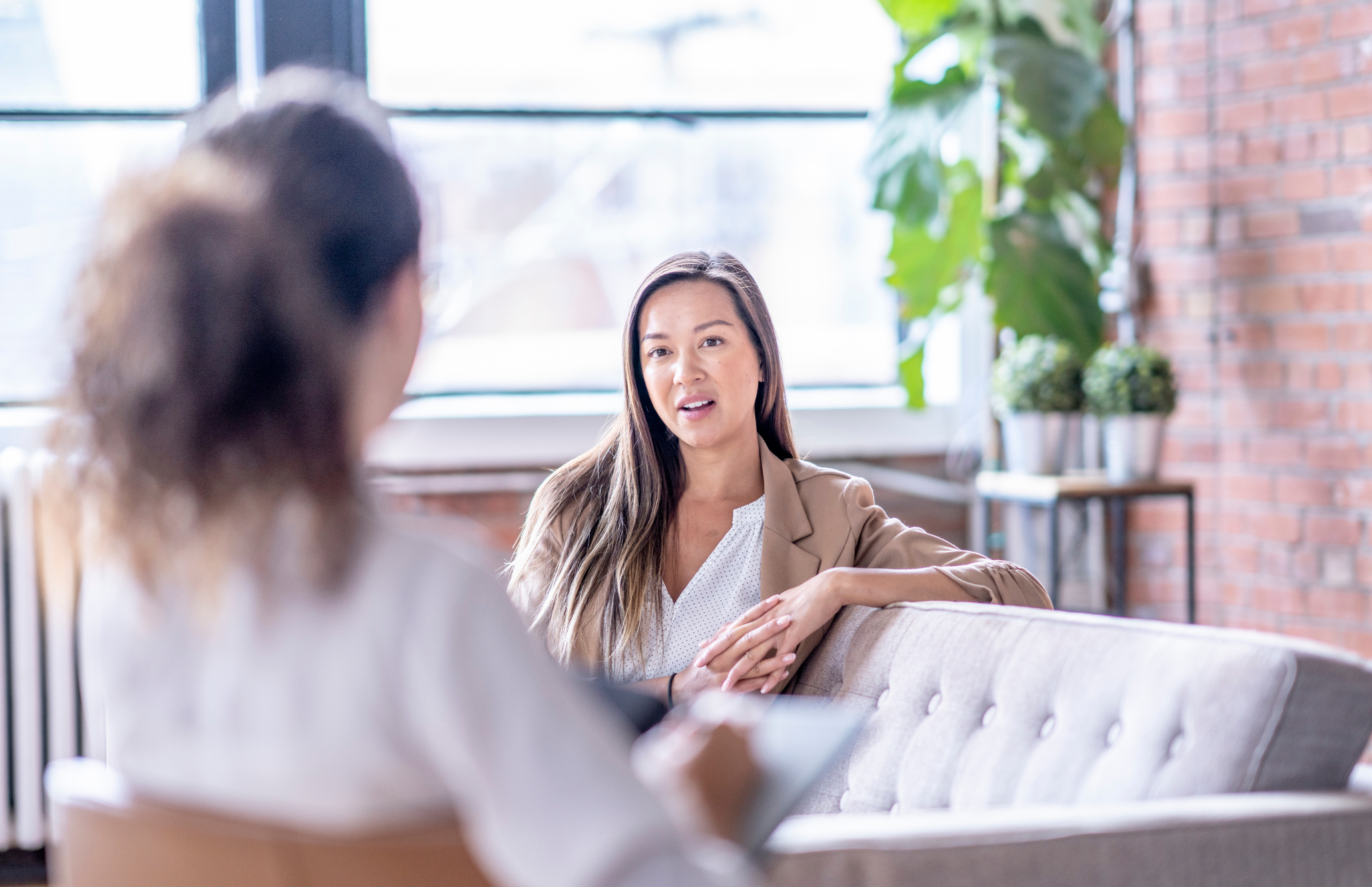 Two women having a conversation in a bright, modern indoor setting with large windows and potted plants.