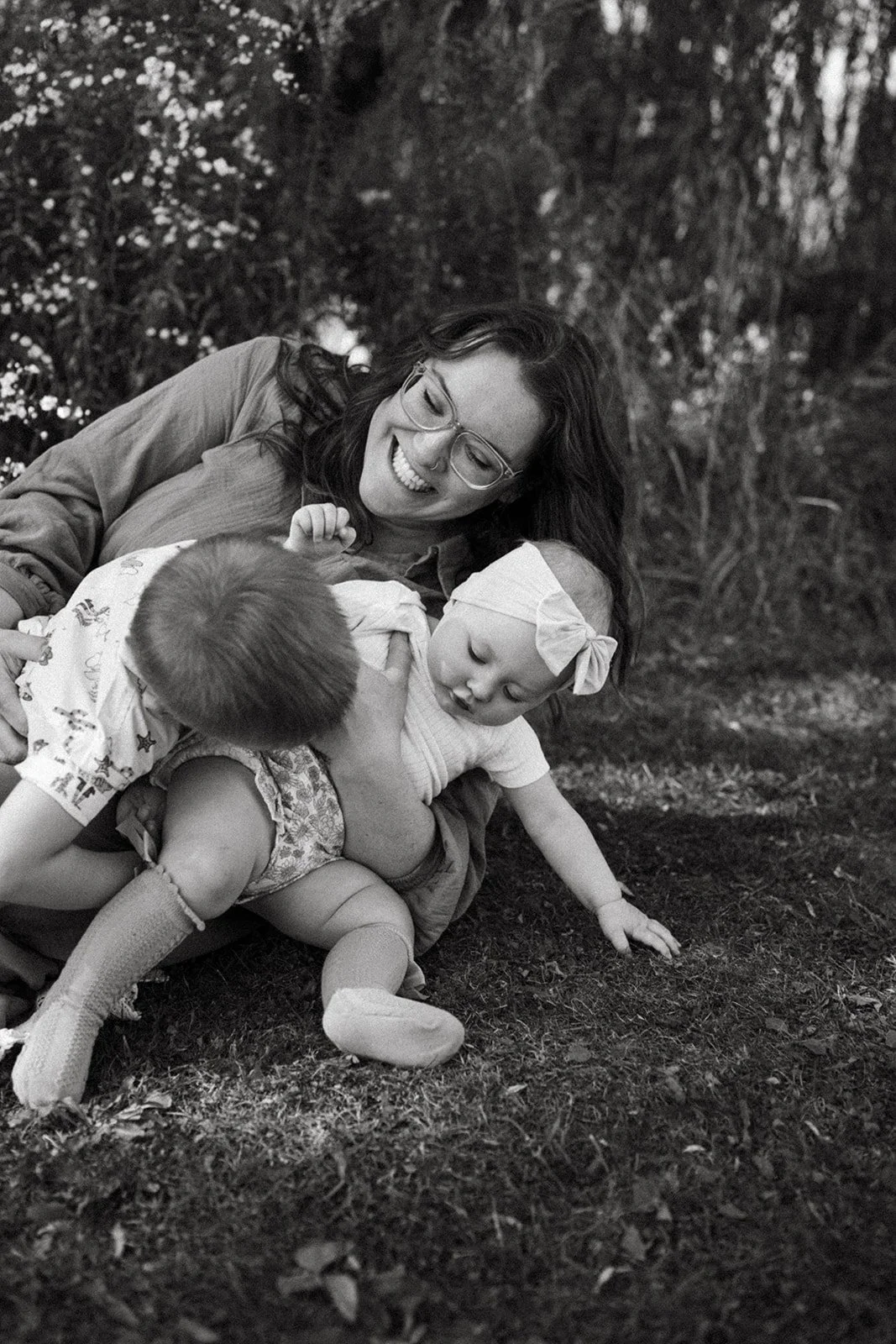 A woman playing with two young children outdoors in a grassy area with trees in the background, captured in black and white.