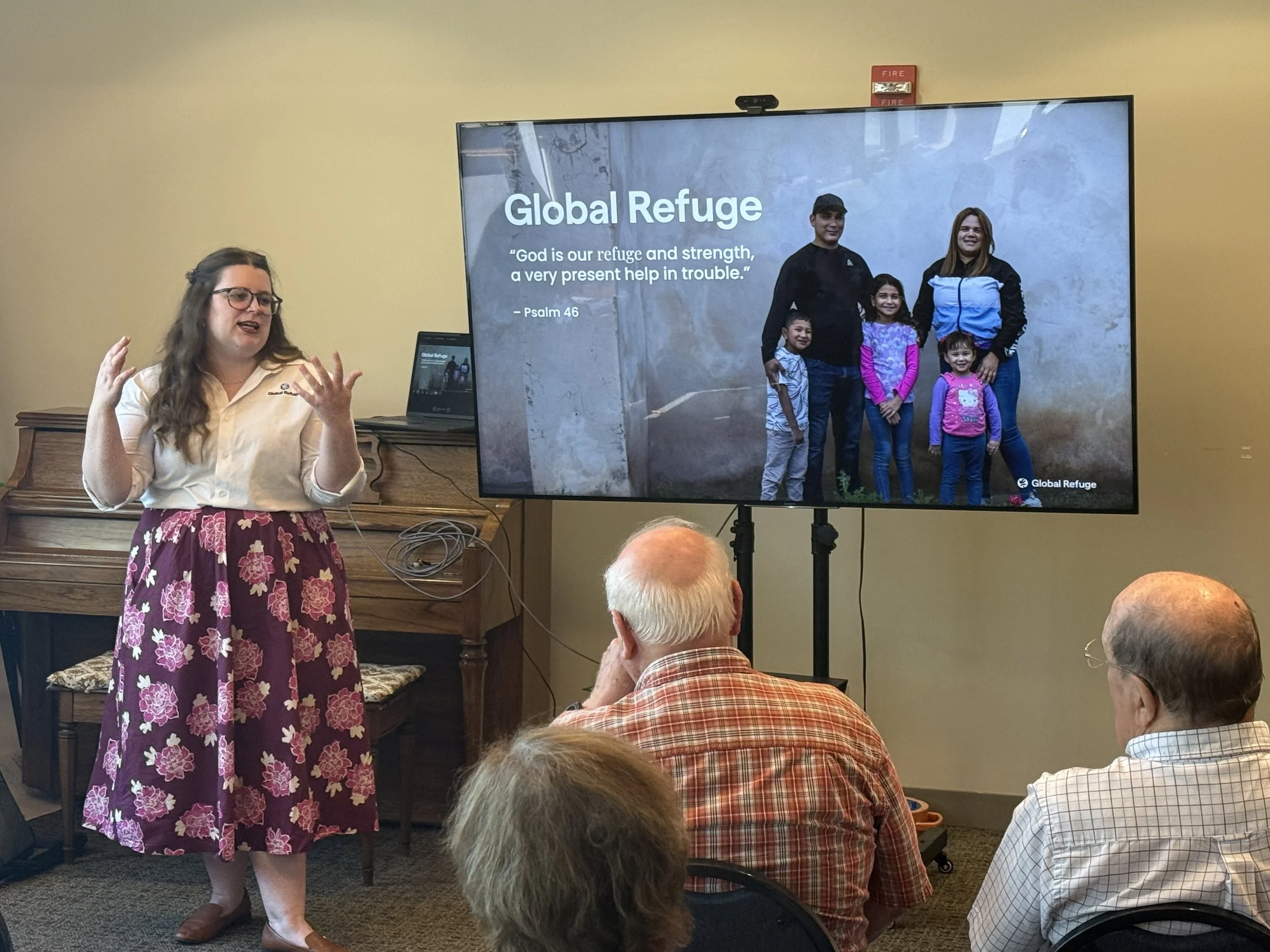 A women speaking to a group of adults next to a screen with an image of a family that reads Global Refuge and refers to Psalm 46