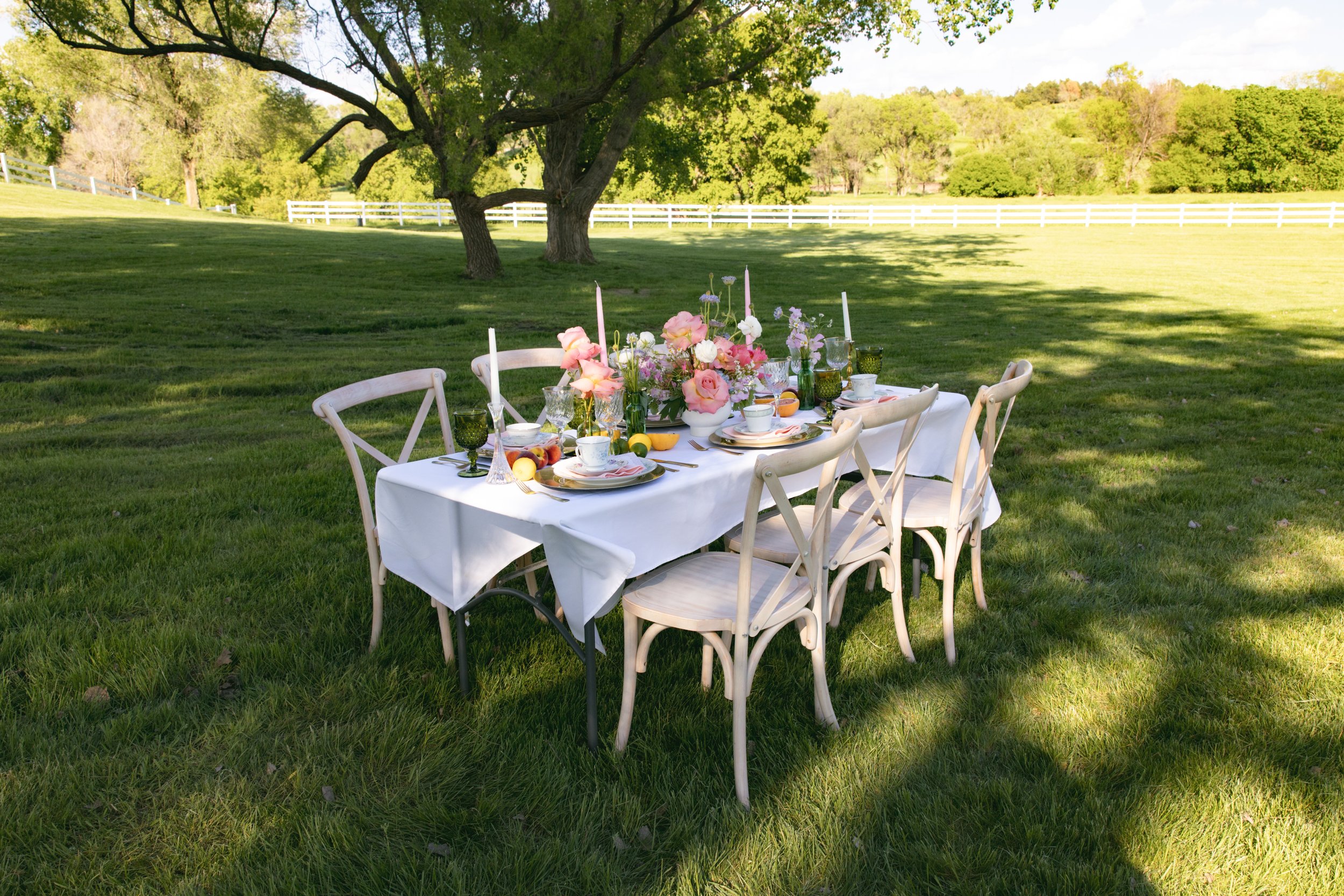 Styled tablescape with white table linen, florals, and serverware located outside near tree with