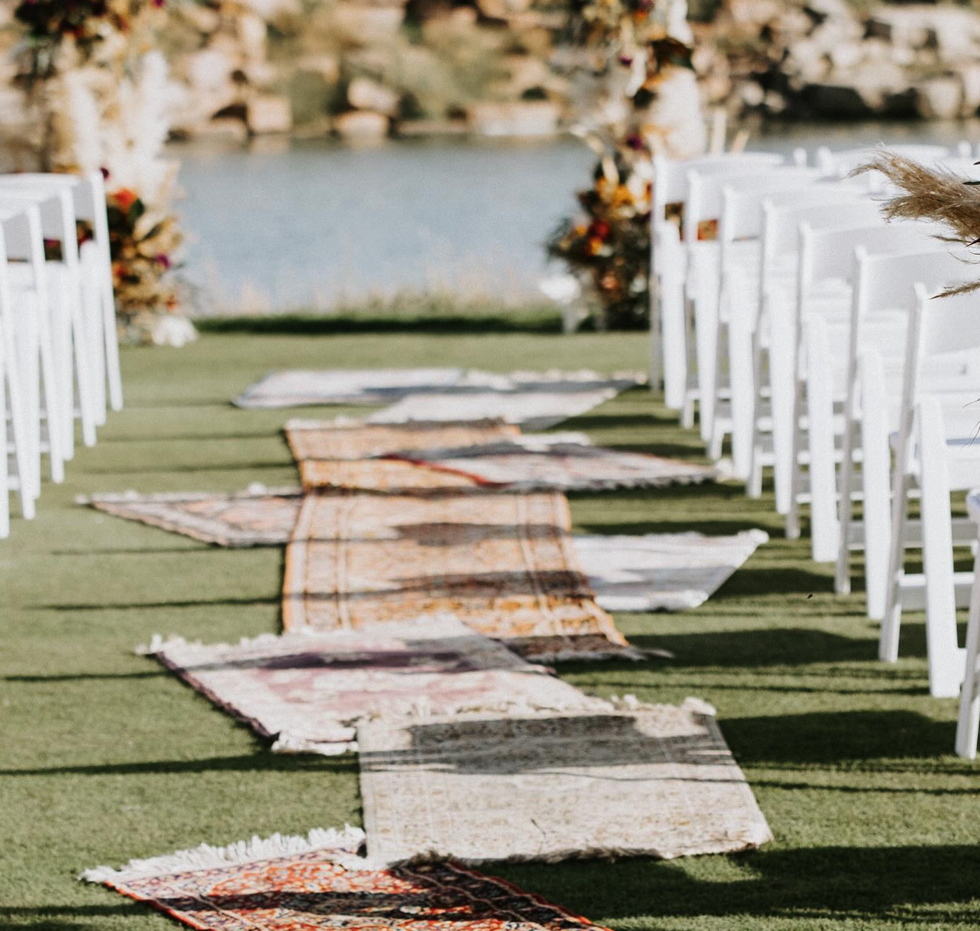 Wedding ceremony aisle outdoors with white chairs and overlapping oriental rugs with a lake in the background