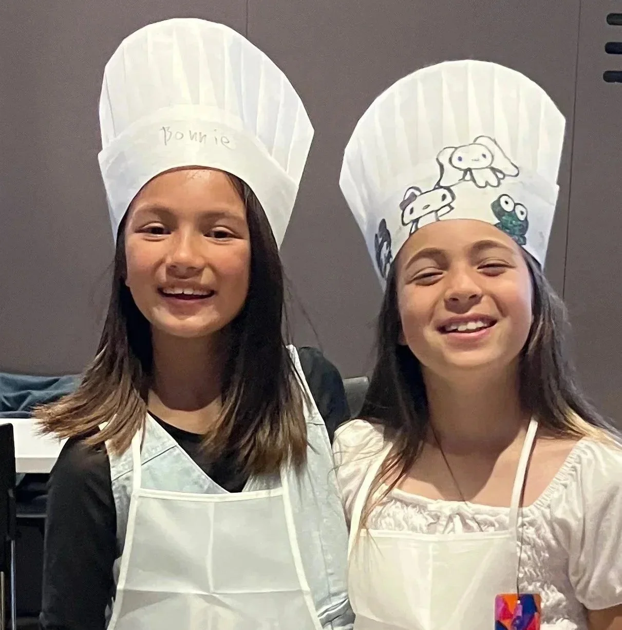 Two kids smiling and wearing chef hats and aprons