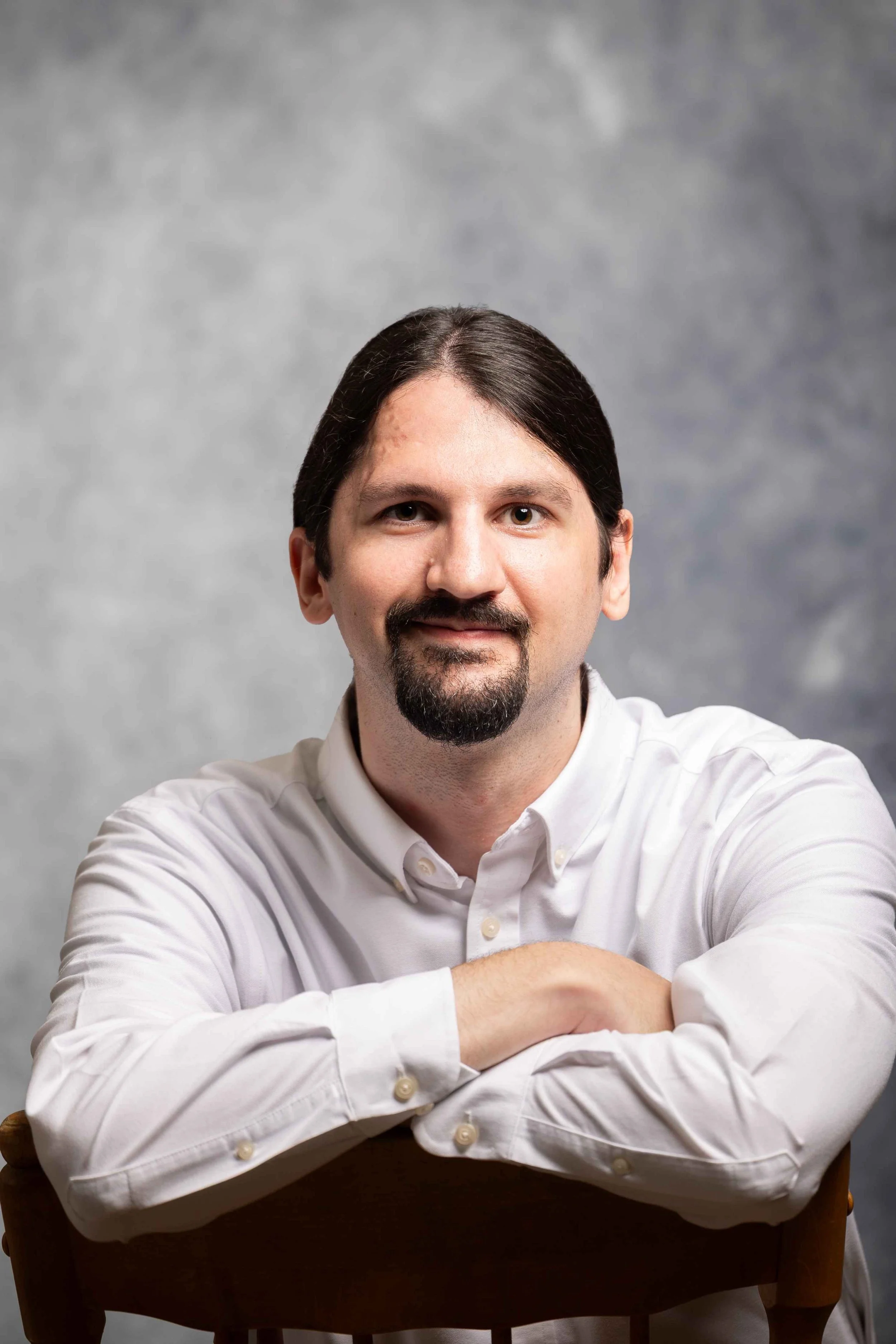 Portrait of a man with dark hair, facial hair, wearing a white button-up shirt, sitting with arms crossed on a wooden chair, against a gray background.
