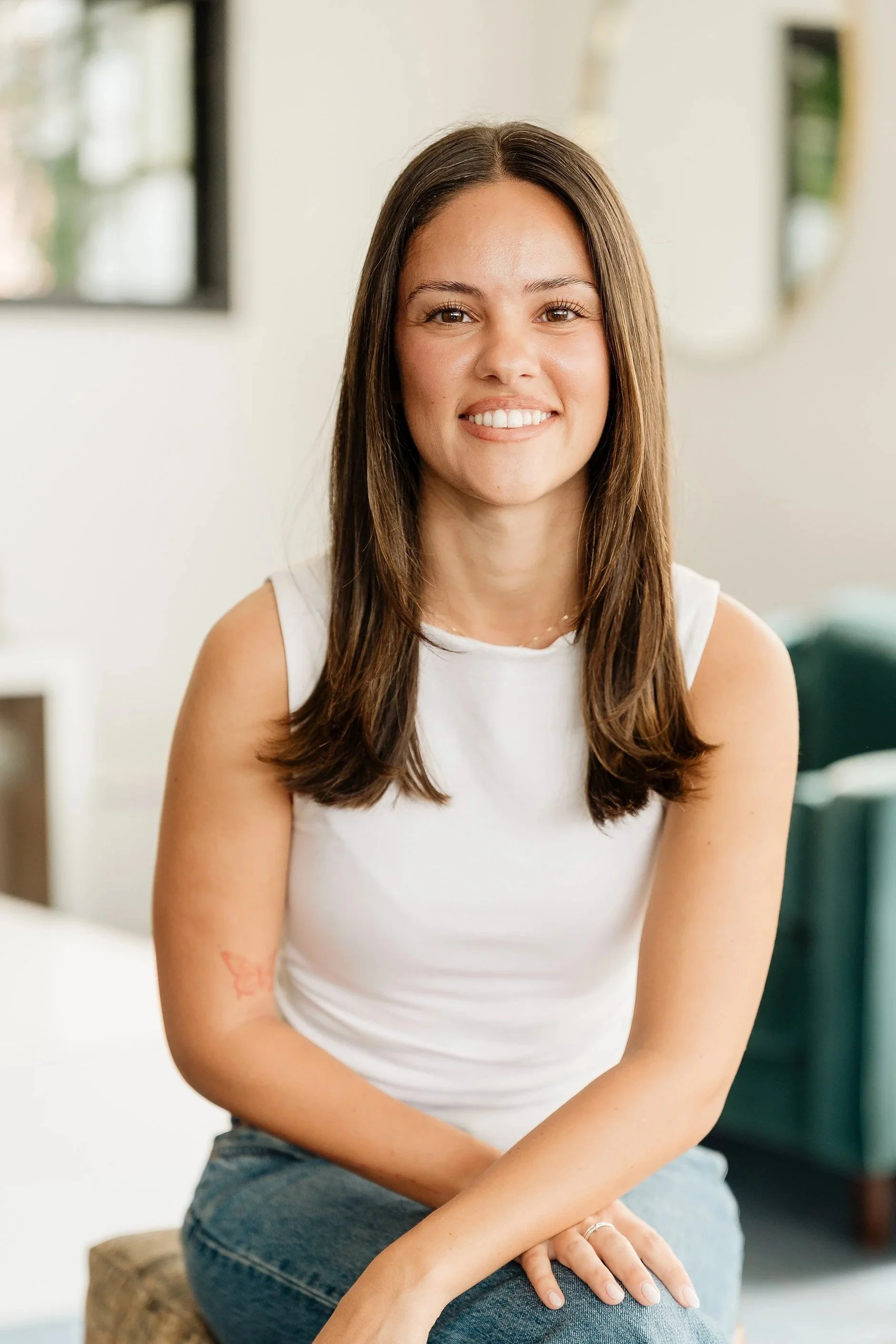 A young woman with long brown hair, smiling, sitting indoors on a wooden stool, wearing a white sleeveless top and blue jeans.