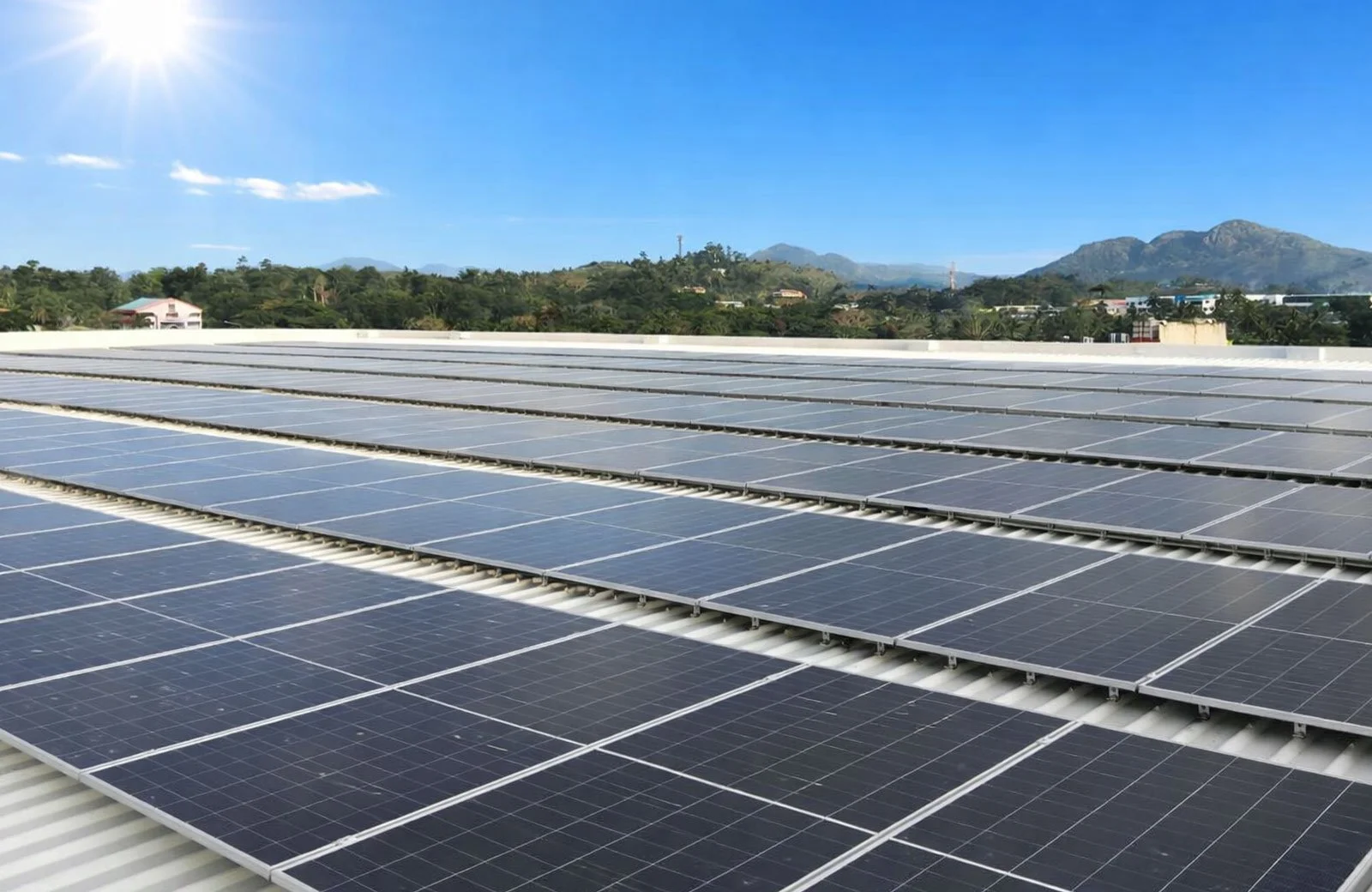 Rows of solar panels on a rooftop with tropical vegetation and mountains under a bright blue sky