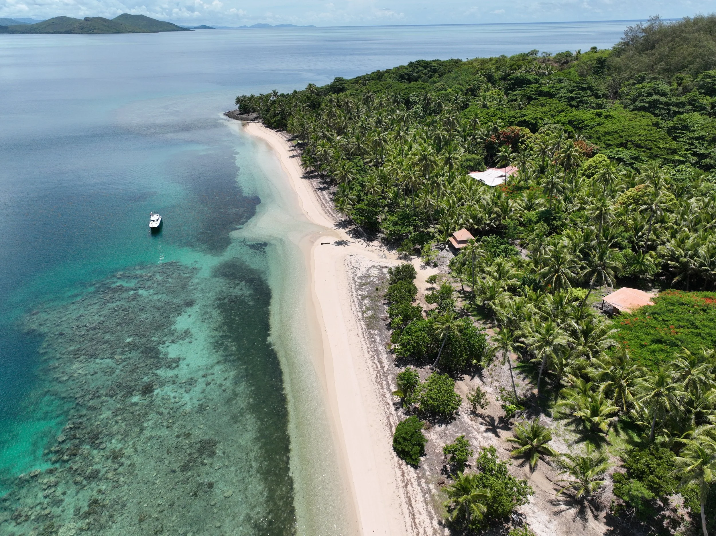 Aerial view of a tropical beach with clear turquoise water, coral reefs, palm trees, small huts, and a boat offshore.
