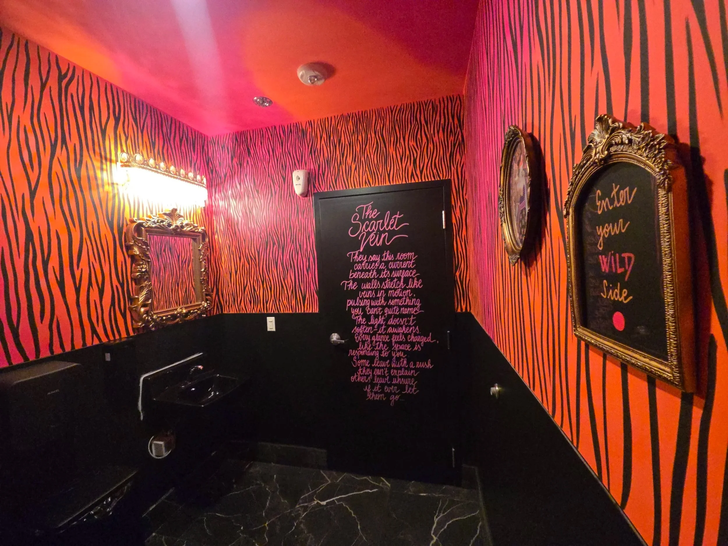 Wide view of The Scarlet Vein Bathroom, featuring hand-painted neon pink and neon orange zebra patterned walls, ornate gold frames, and immersive interior details by Pao Saenz at 4 Dreams Escape Rooms & Lounge in Redmond, Washington.