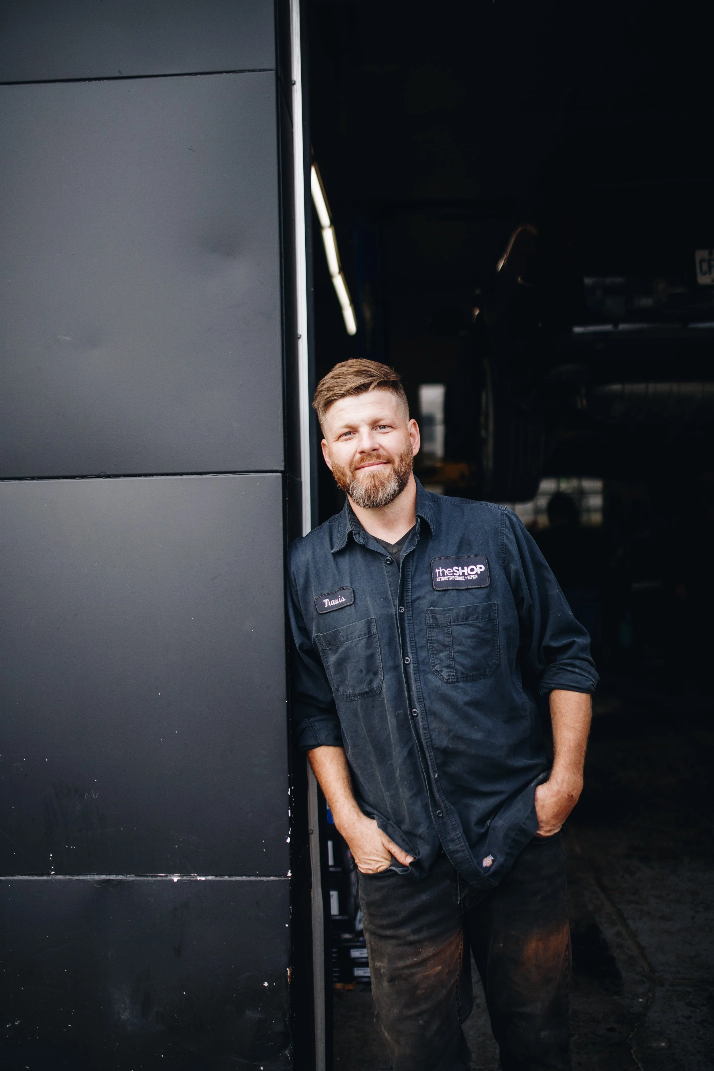 A man with brown hair and a beard, wearing a black work uniform with a name tag that says Tony, stands with his hands in his pockets, leaning against a black partition in an automotive repair shop.