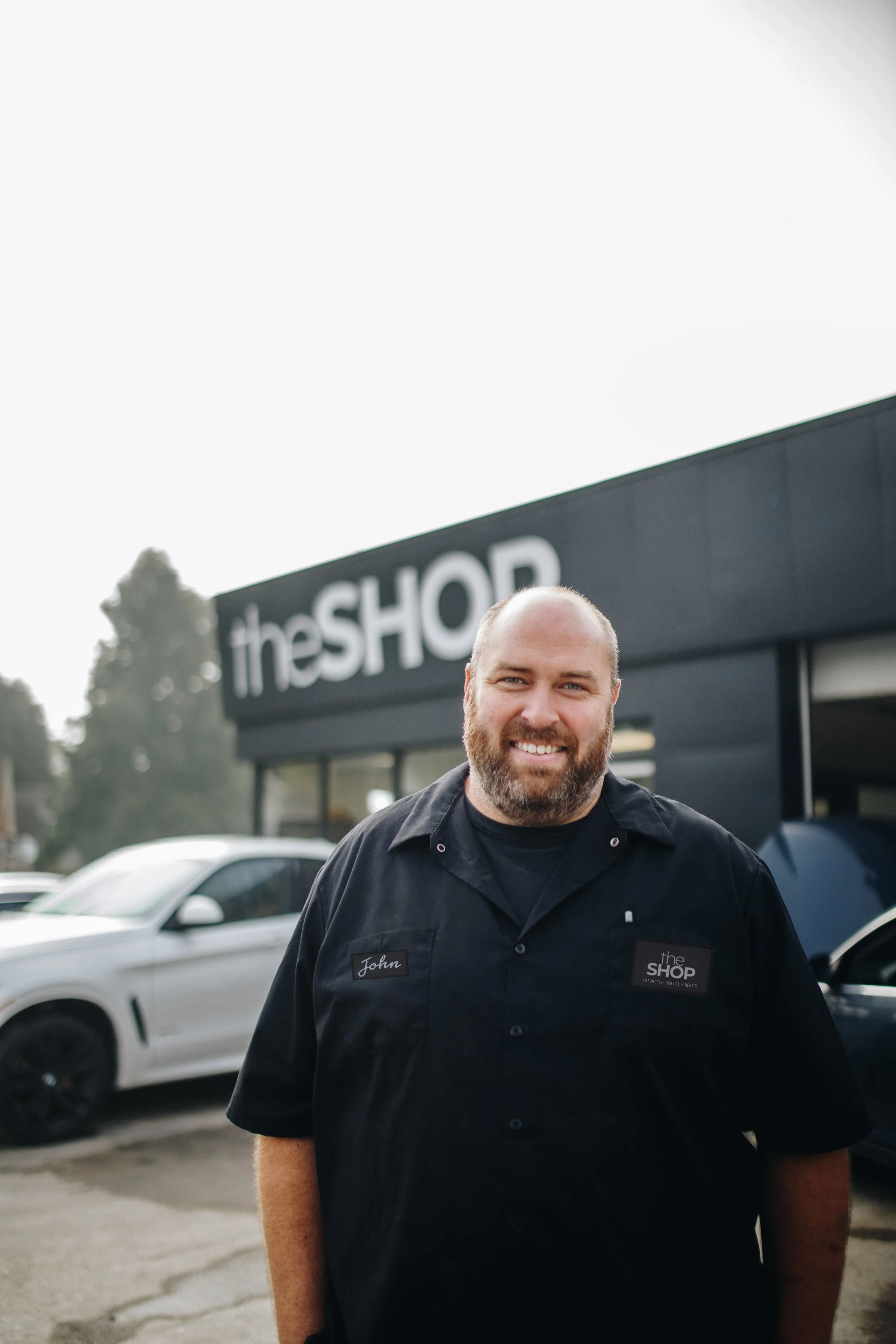 A man with a beard and a black uniform standing in front of a storefront with the sign 'theSHOP' visible behind him, surrounded by cars in a parking lot.