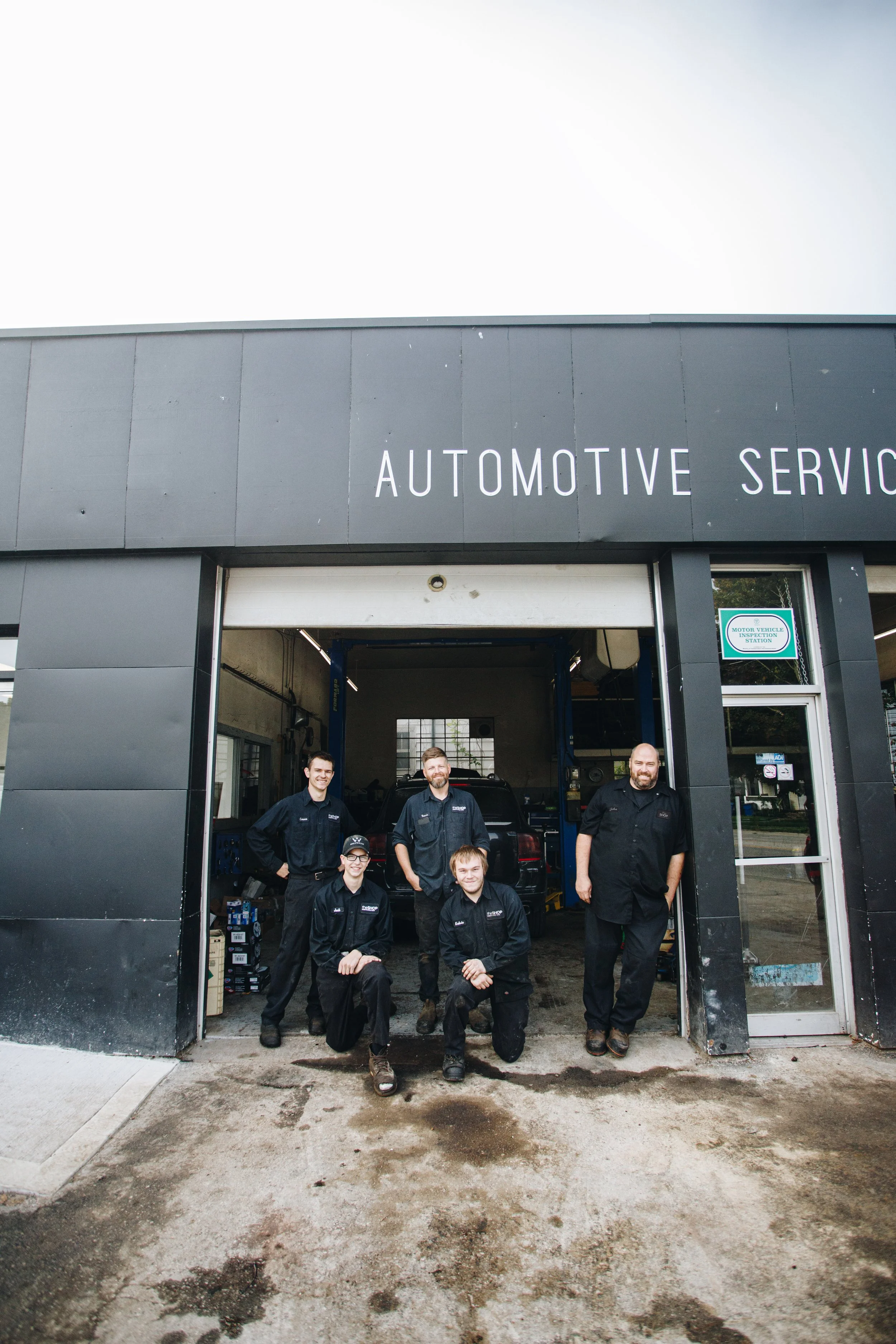 Group of five auto service workers standing outside a garage, smiling for the photo, with some kneeling and others standing behind them.