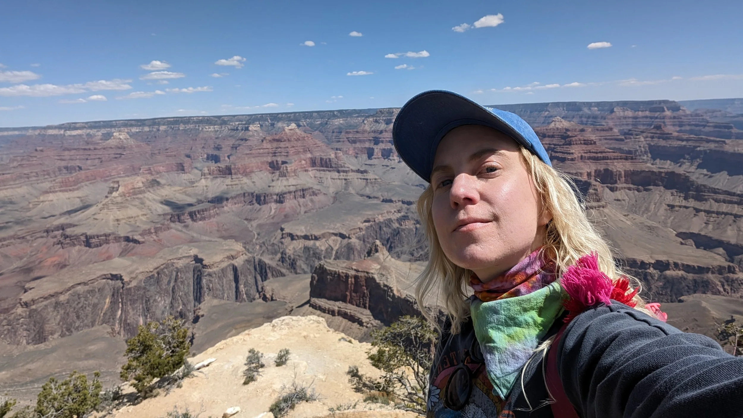 Person taking a selfie at the Grand Canyon under a clear blue sky with scattered clouds.