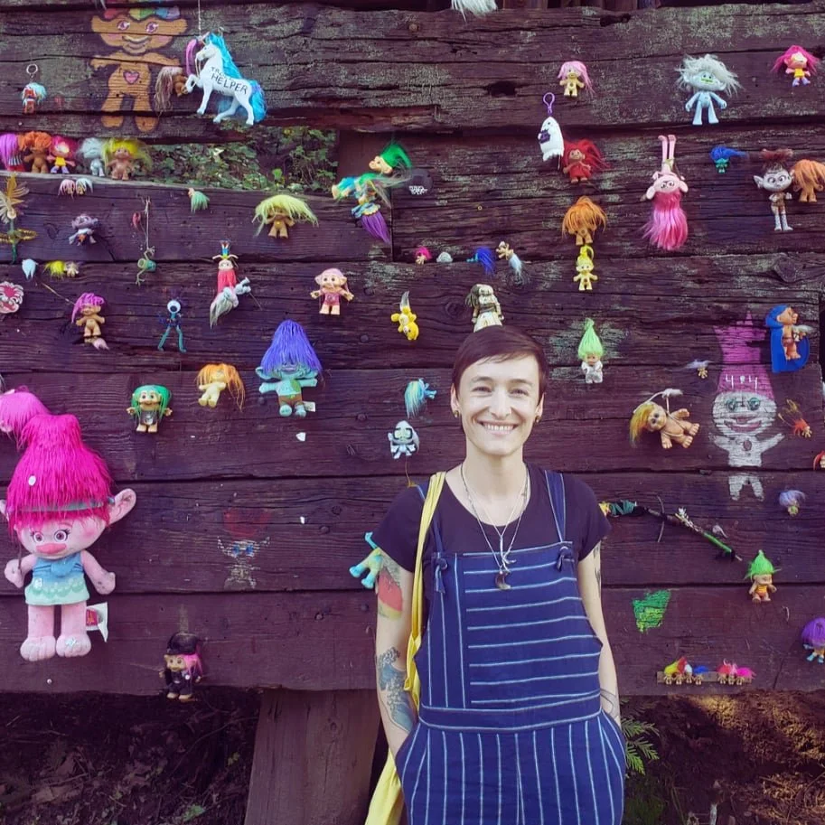 Sarah, with cropped hair, wears a blue jumper and stands before a bridge with multicolored trolls affixed to it.