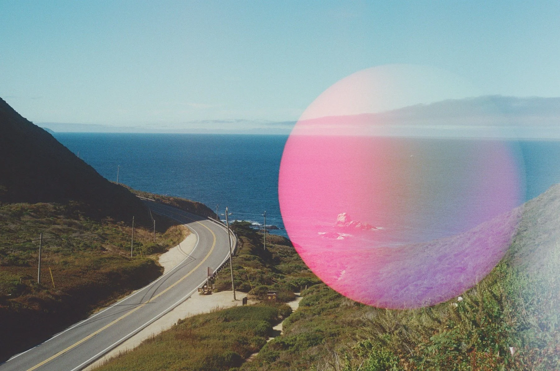A film photo of a winding California highway, with the Pacific Ocean in the background and a large pink light leak circle on the right half of the frame.