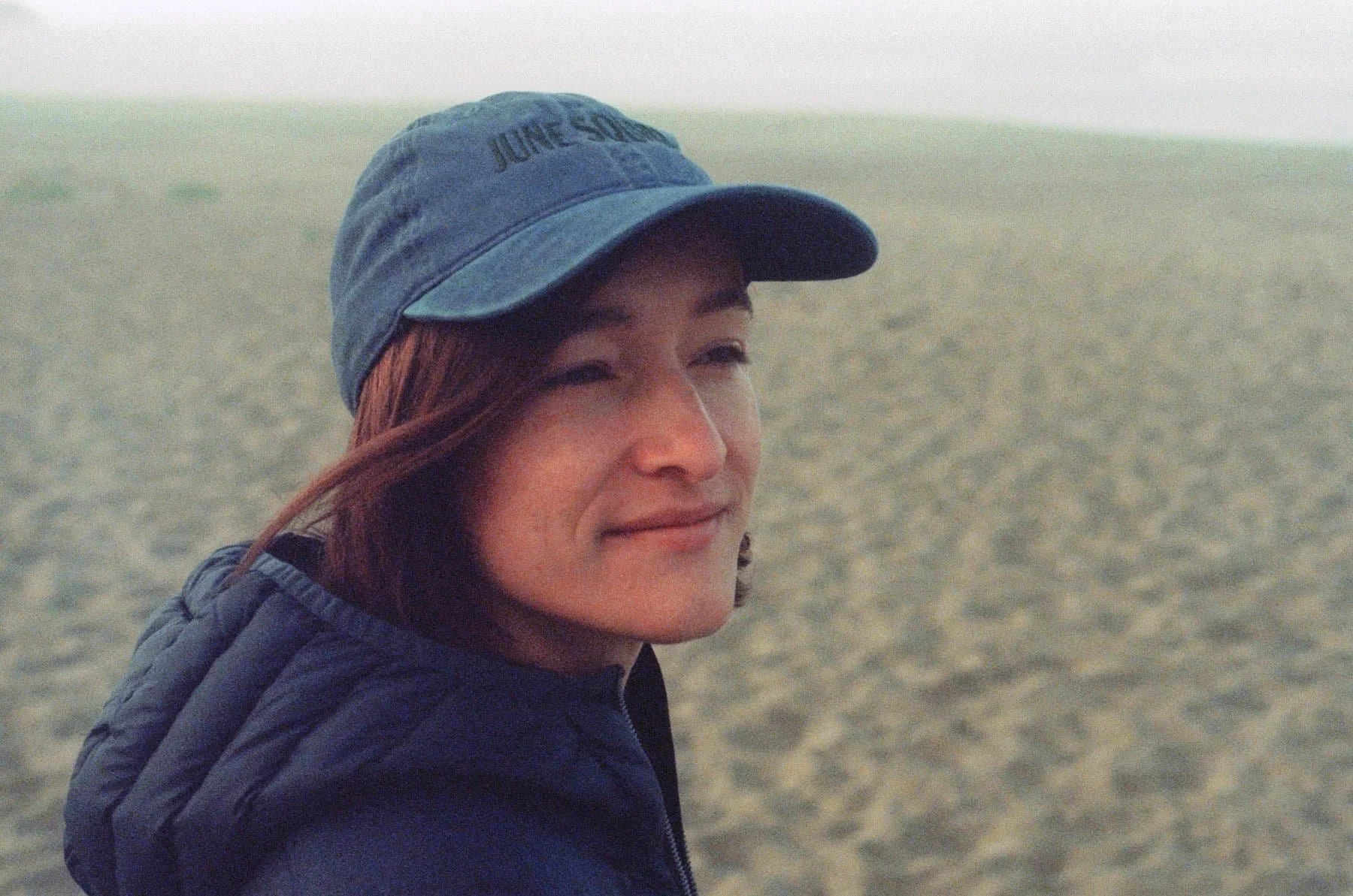 Sarah wears a blue hat and jacket while standing at the beach, looking to the right of the frame.