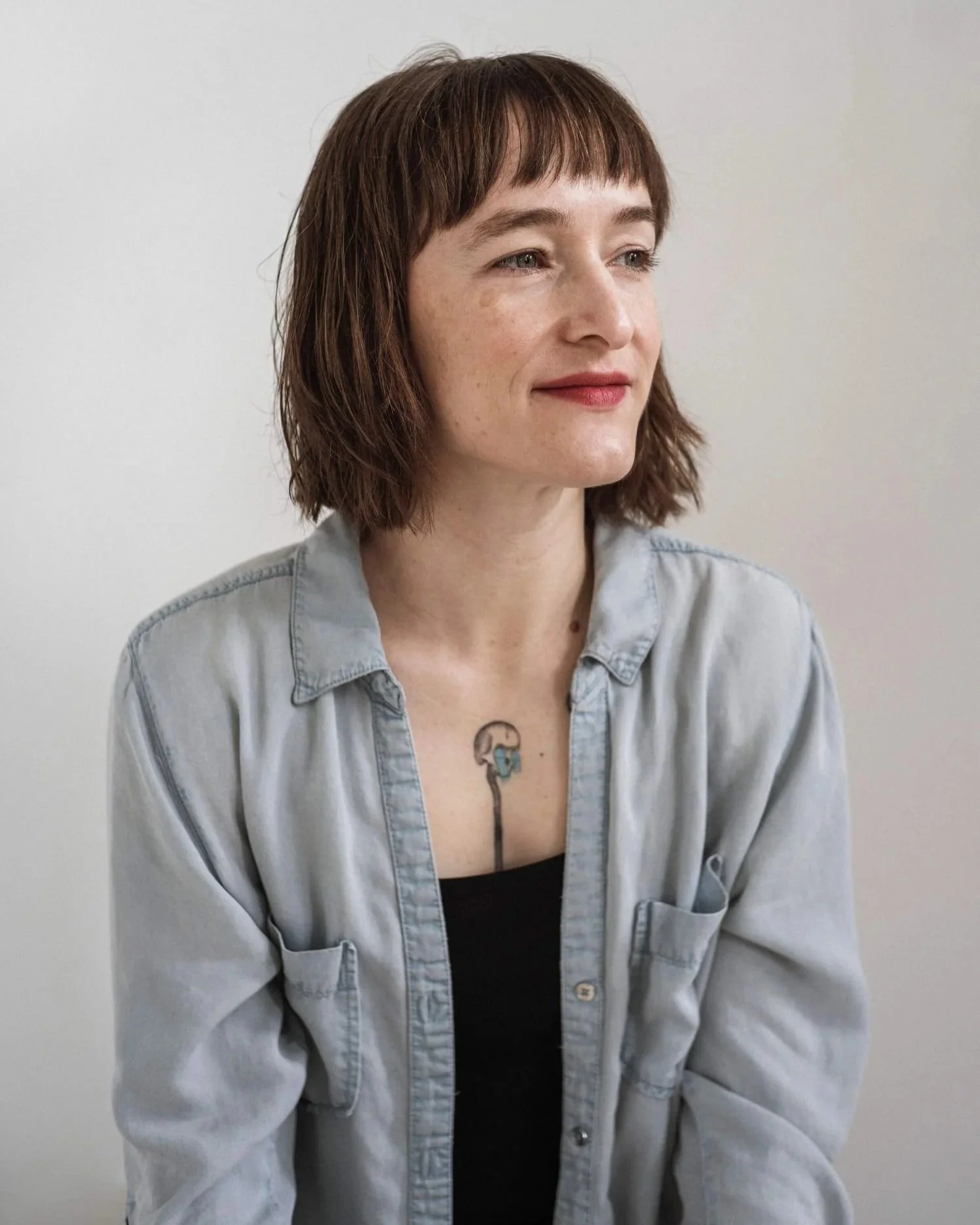 A woman with short brown hair, light skin, and freckles sitting against a plain light background, wearing a light-colored button-up shirt over a black top, with a colorful tattoo of a flamingo on her chest.
