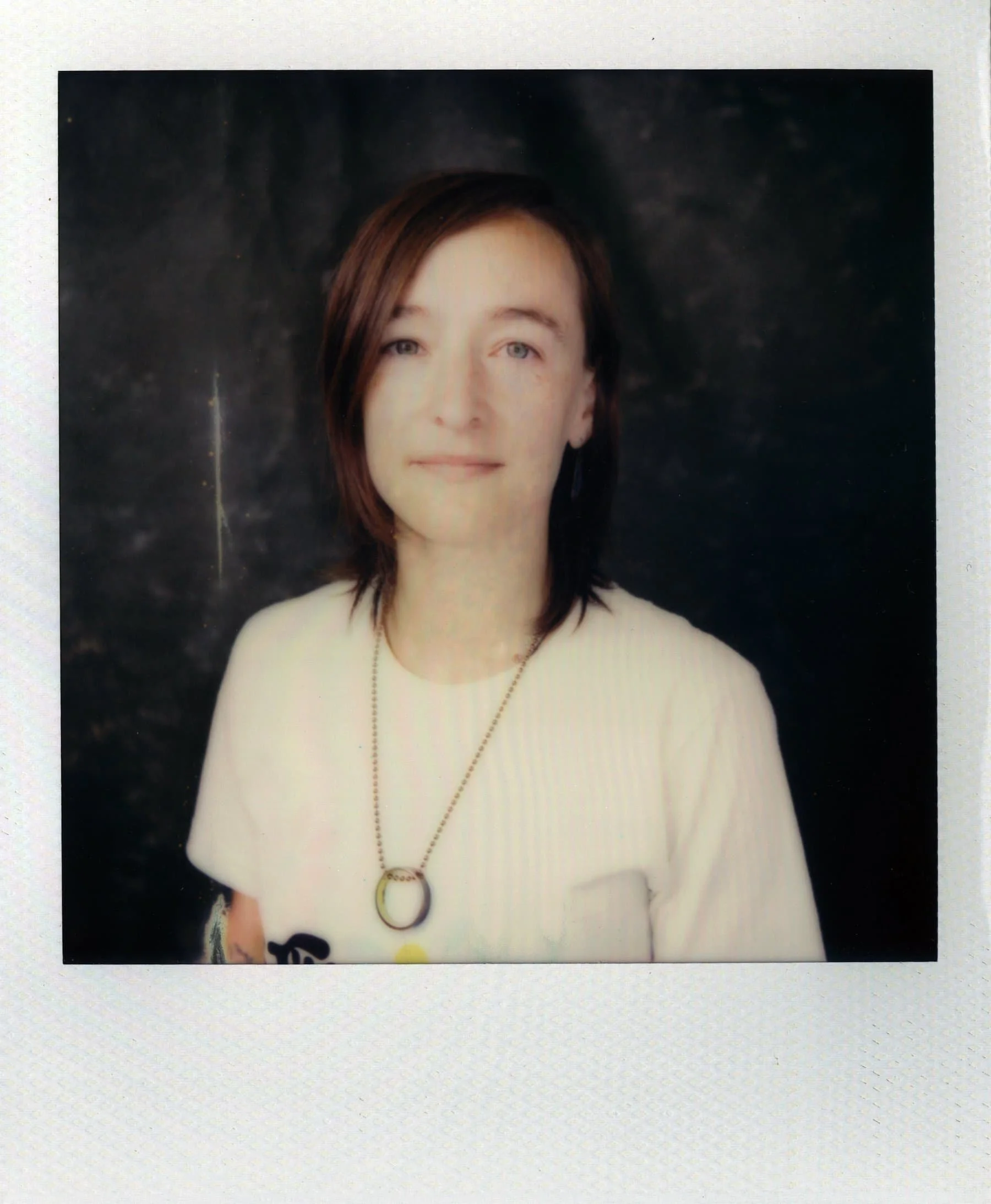 A Polaroid portrait shows Sarah wearing a beige shirt and a long necklace with a golden circle at the end. Her brown hair is shoulder length, and she looks directly at the camera.
