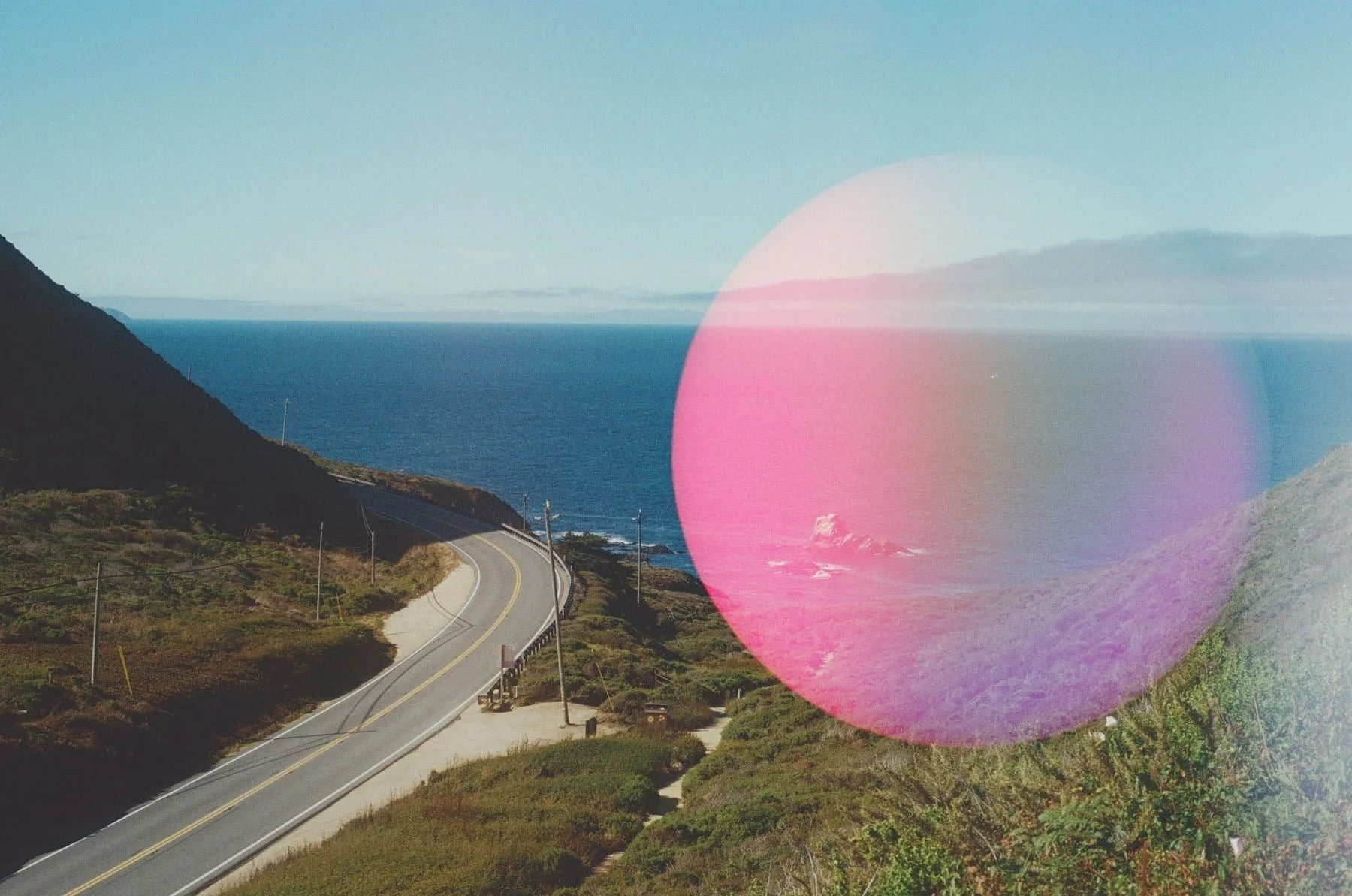 A film photo of a winding California highway, with the Pacific Ocean in the background and a large pink light leak circle on the right half of the frame.