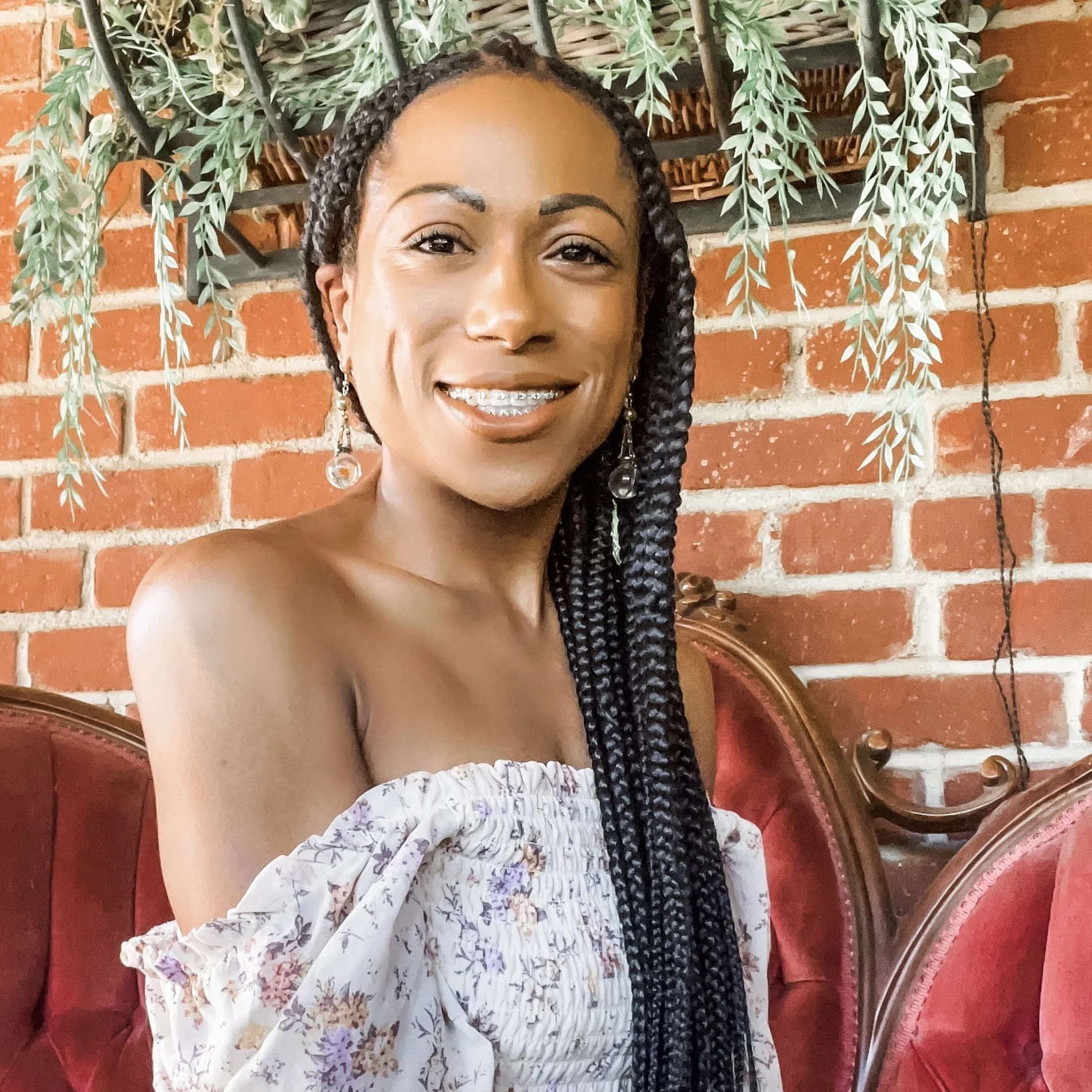 A woman with braided hair and earrings smiling in front of a brick wall decorated with hanging leafy plants and a jewelry stand.