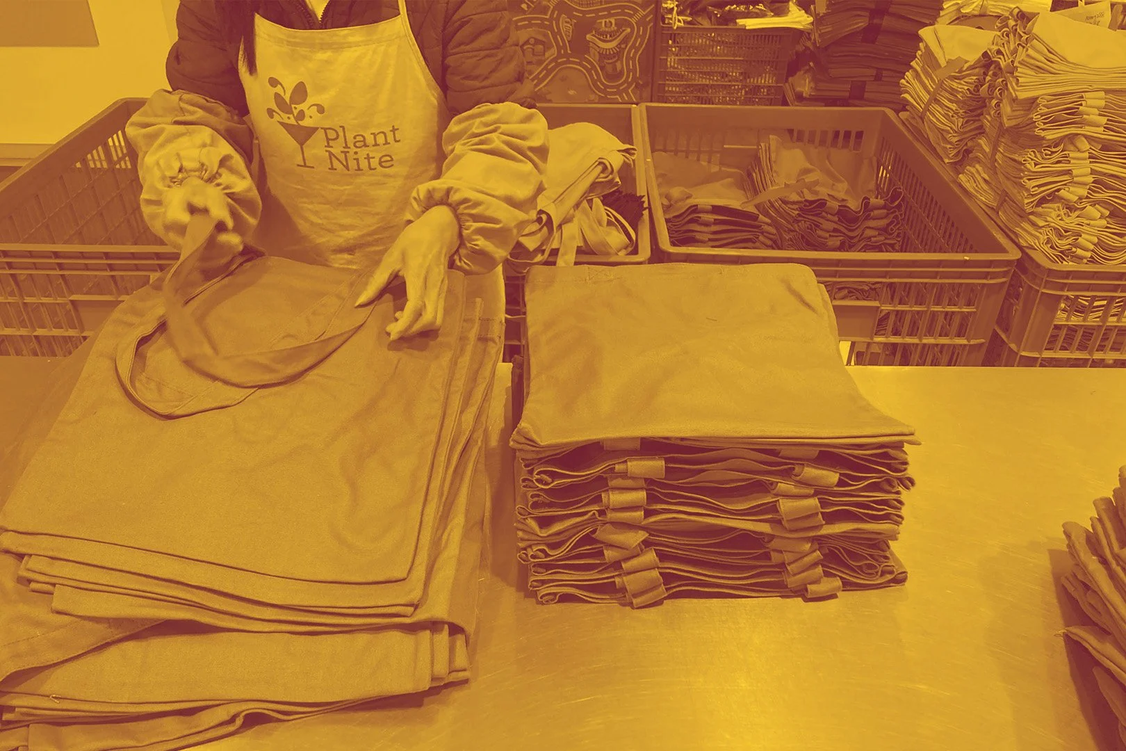 A person's torso wearing a white apron labeled 'Plant Nite' stands behind a table with folded linen napkins and cloths; reusing a shade of yellow.